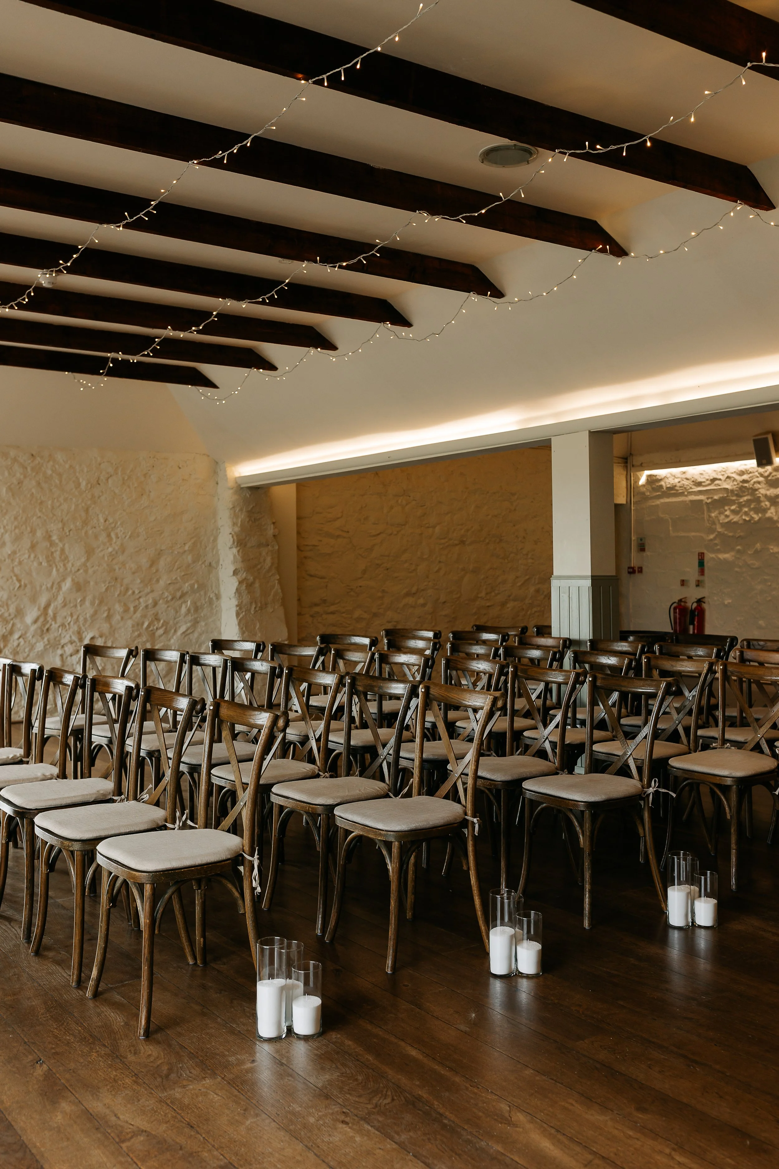 Empty chairs arranged in rows inside a decorated event space with candles on the floor and string lights hanging from the ceiling beams.