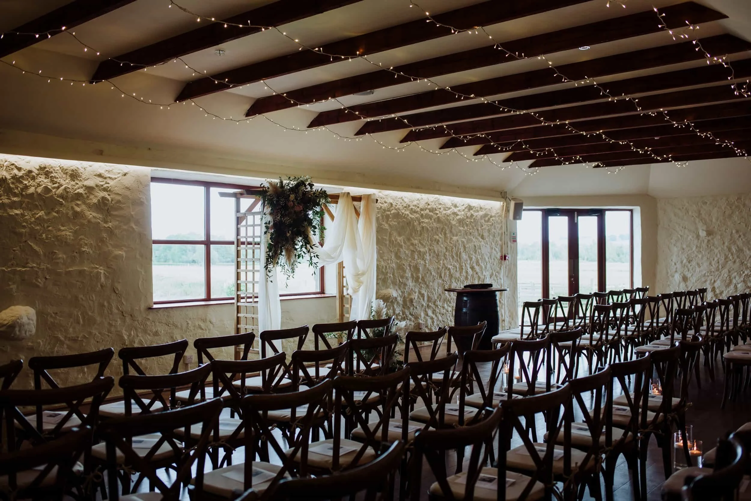 Indoor wedding ceremony setup with wooden chairs arranged in rows facing an arch decorated with flowers and draped fabric, illuminated by string lights on a wooden-beamed ceiling, with windows and a door showing an outdoor view.