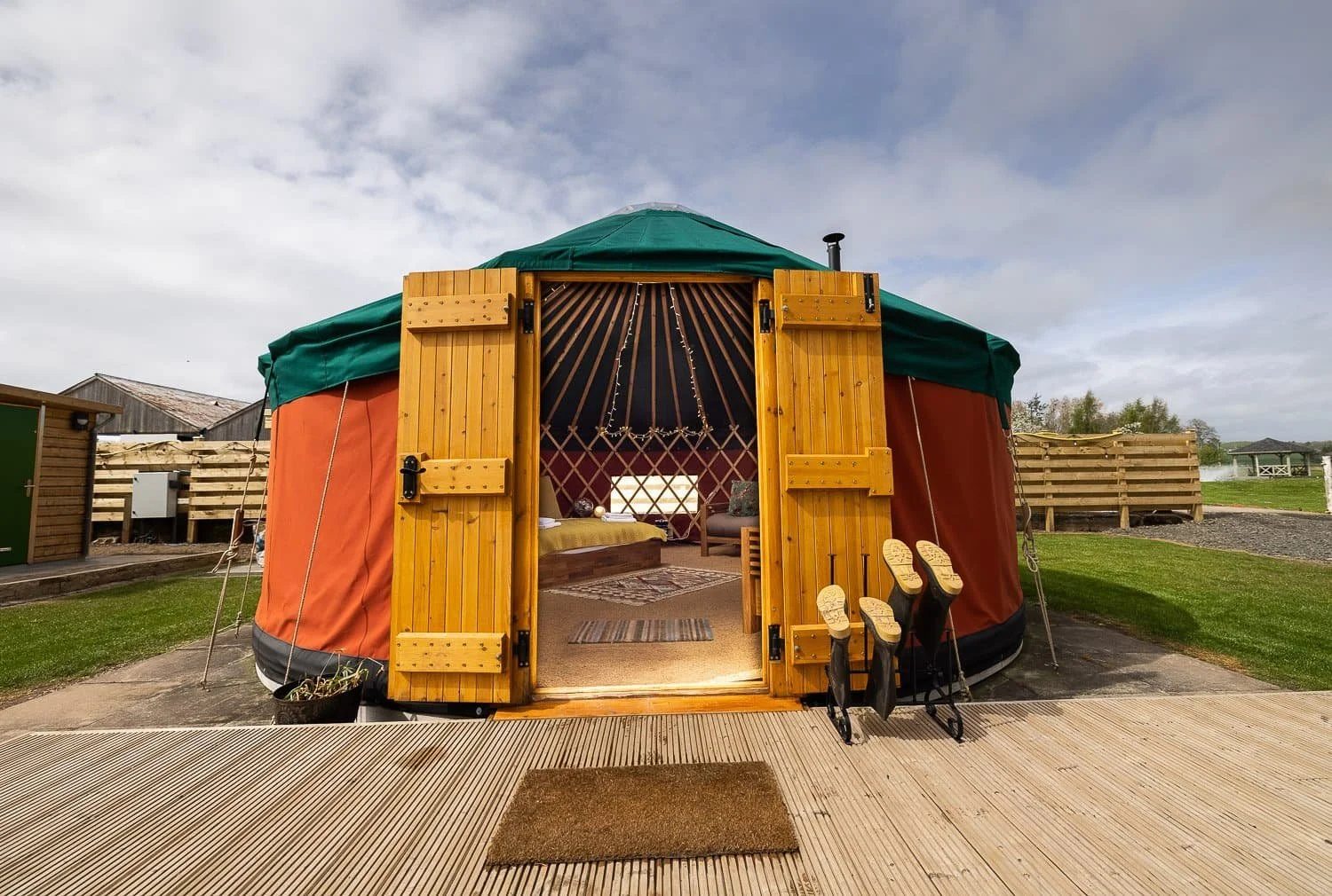 Exterior of a traditional yurt with wooden doors open, showing interior with a bed, rugs, and furniture, set on a wooden deck in a grassy area under a cloudy sky.