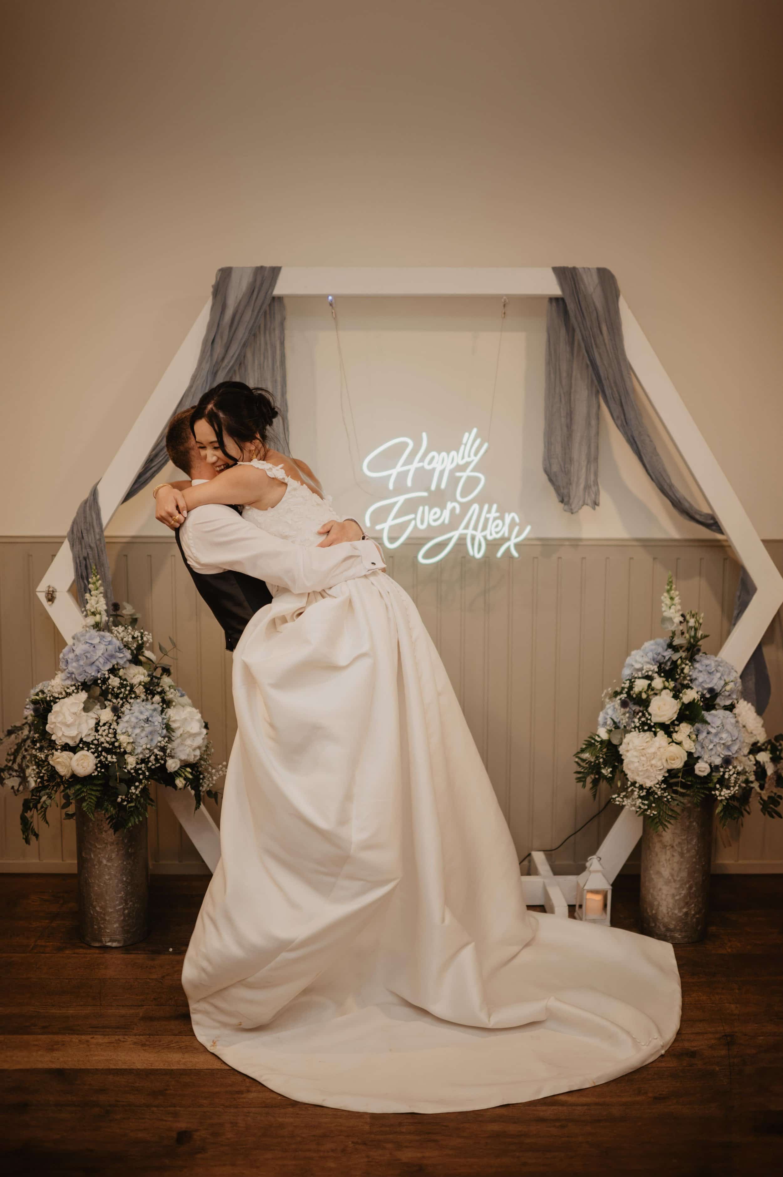 A bride and groom embrace in front of a wedding backdrop with a neon sign that reads 'Happily Ever After.' The bride is wearing a white wedding gown, and the groom is in a black vest and white shirt. The scene is decorated with white and light blue f