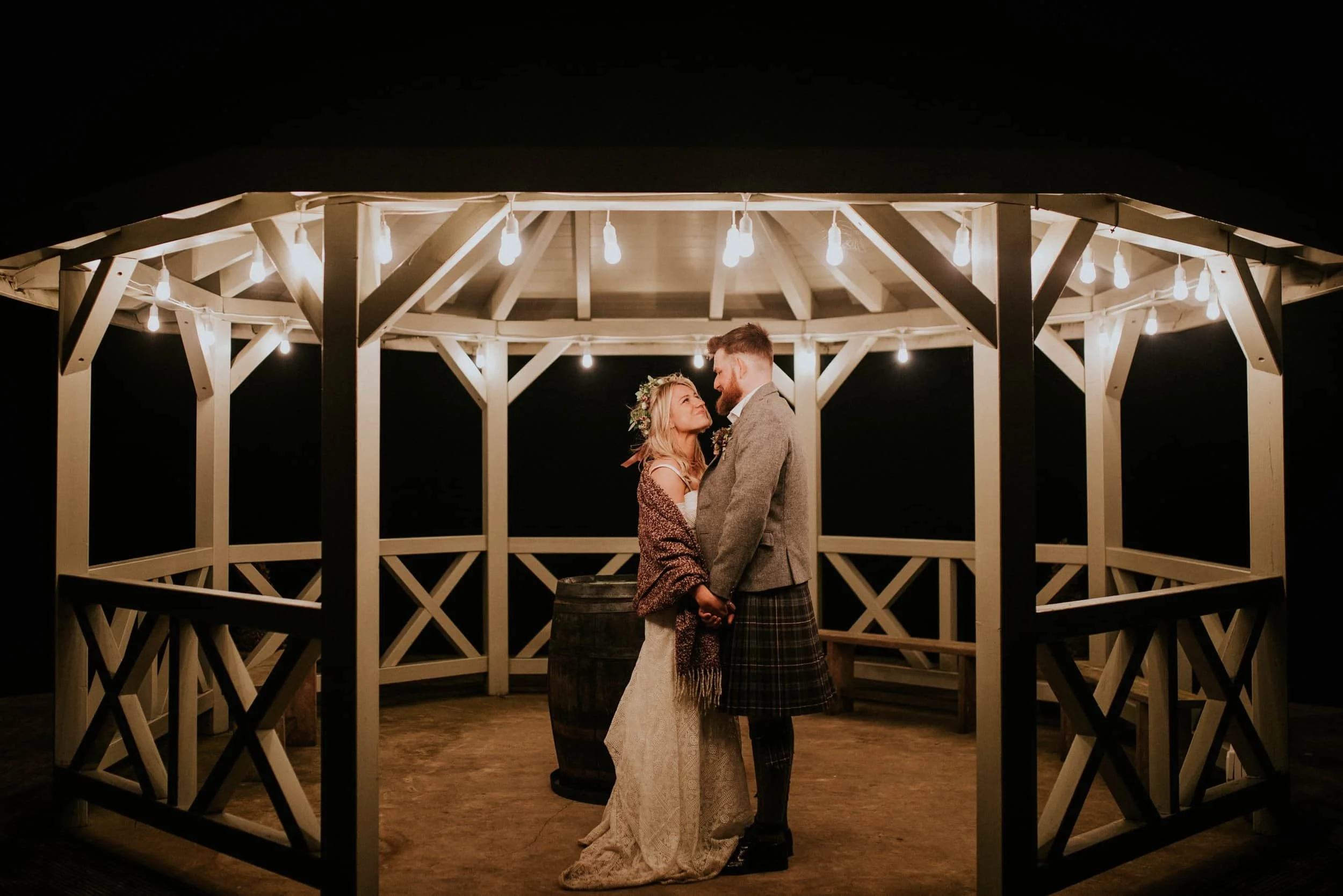 A couple in wedding attire standing closely together under a gazebo decorated with string lights at night.