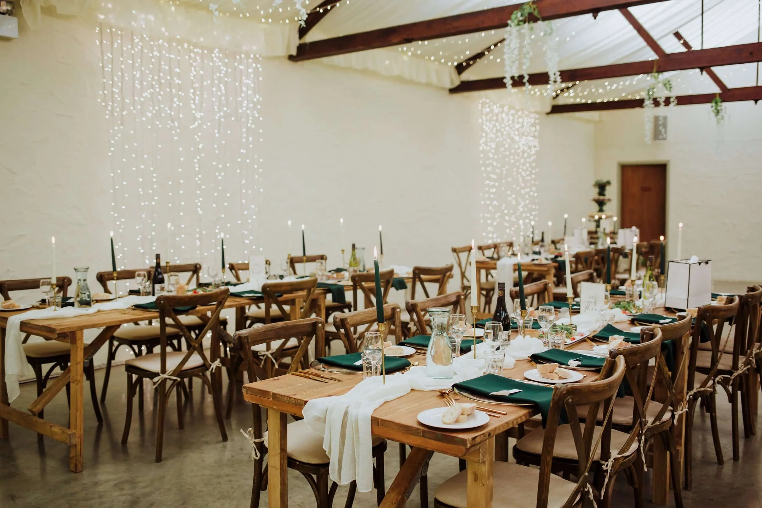 Wedding or event reception setup with long wooden tables, chairs, candle holders, and tableware in a rustic-style hall decorated with hanging string lights and greenery.