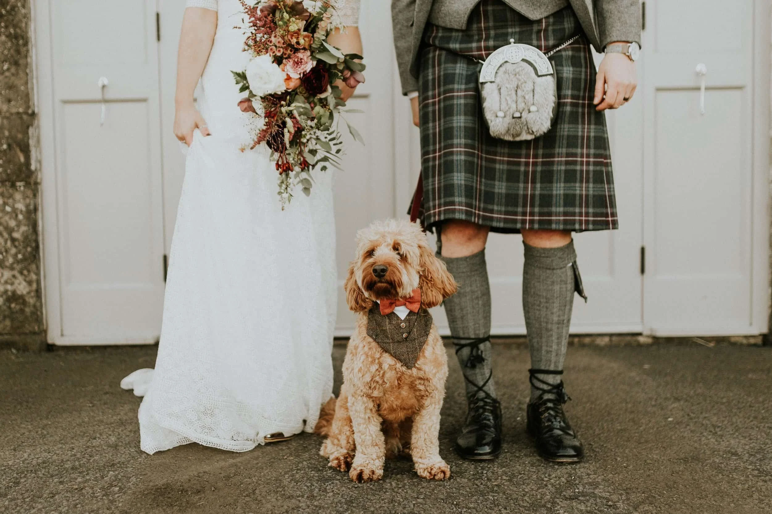 A groom and bride with a dog dressed in a bow tie and vest, standing in front of a white door at a wedding. The bride is holding a bouquet of flowers, and the groom is wearing traditional Scottish attire including a kilt and sporran.