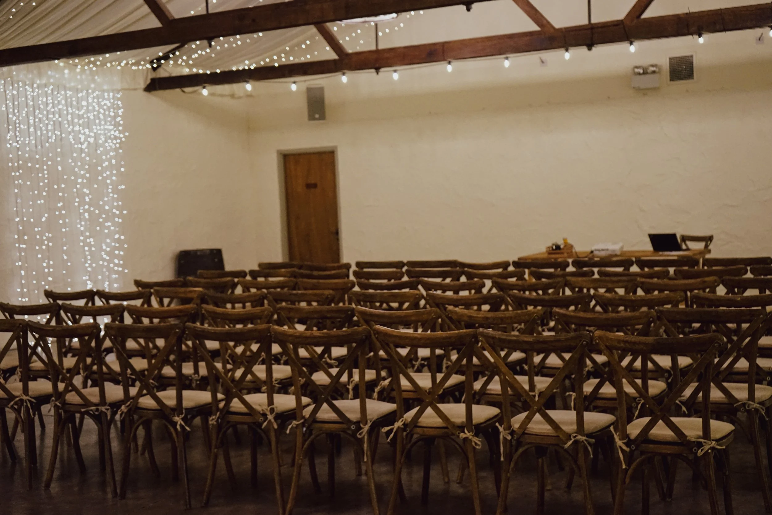 Interior of a hall decorated with string lights and wooden beams, arranged with rows of chairs facing a small table at the front.