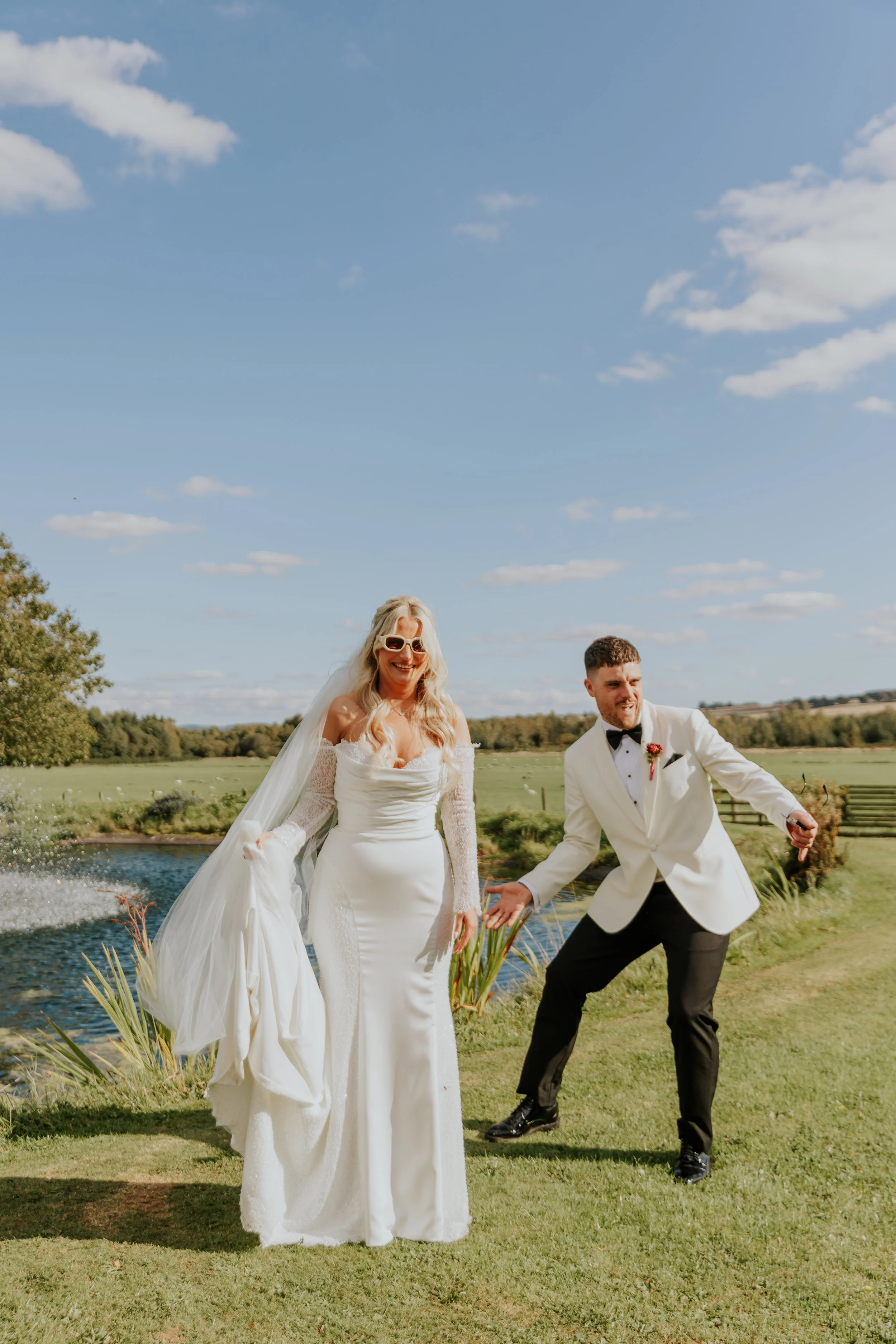 Bride and groom celebrating outdoors beside a pond, smiling and in formal wedding attire, with green grass, trees, and a blue sky with clouds in the background.