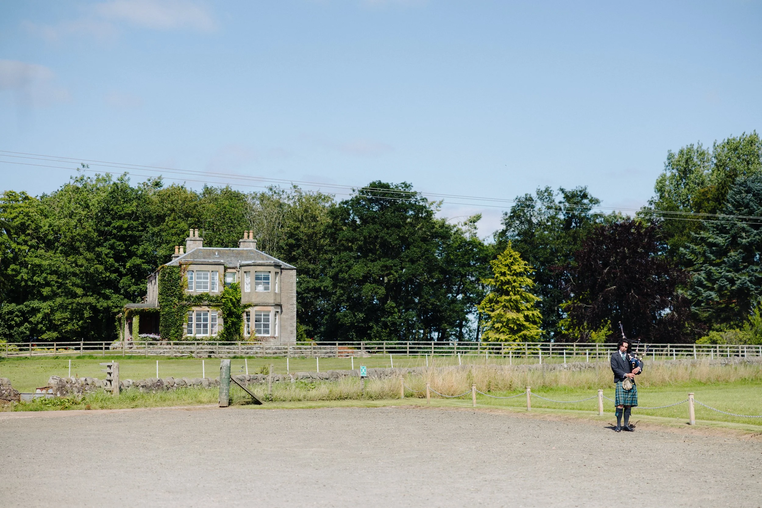 A man dressed in traditional Scottish attire playing bagpipes outdoors near a grassy area with a house and trees in the background.