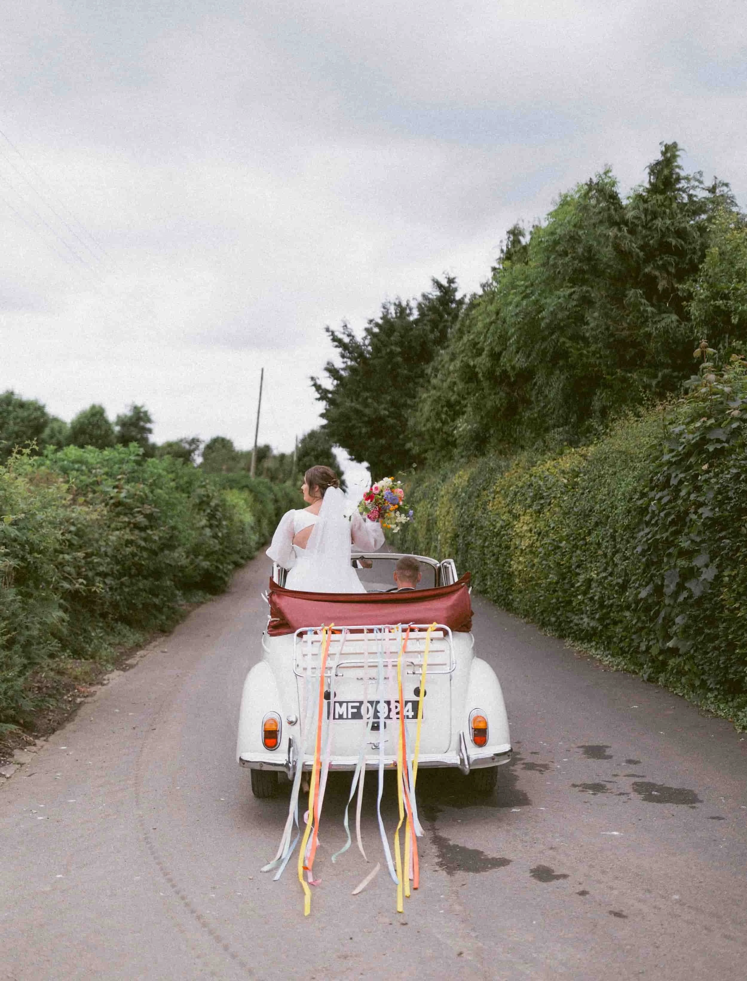 Bride in white wedding dress riding in a vintage convertible car decorated for wedding, holding a bouquet, driving down a narrow road with green foliage on both sides.