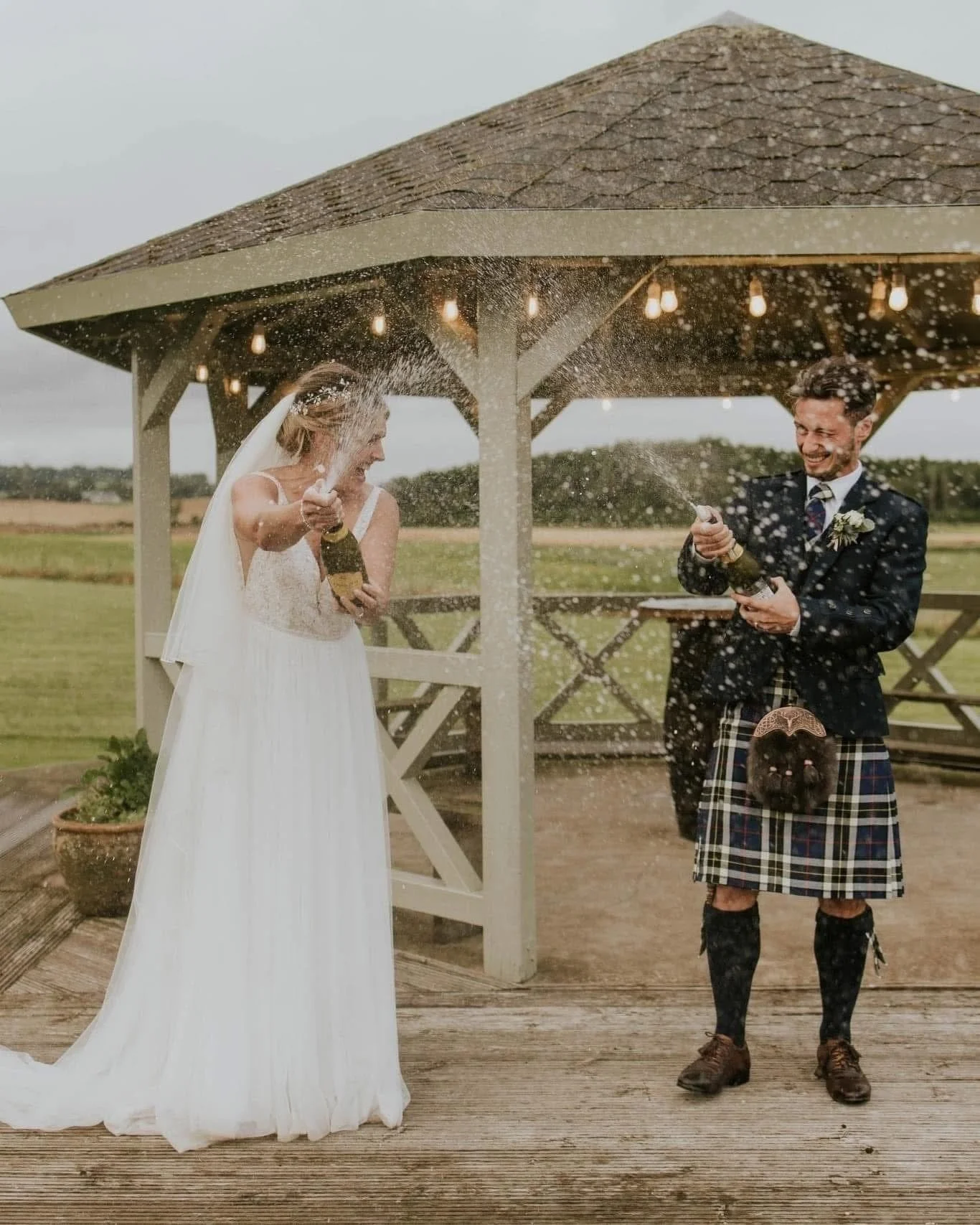 A bride and groom celebrating with champagne under a gazebo outdoors. The couple is dressed in wedding attire, and they are spraying champagne, smiling and laughing.