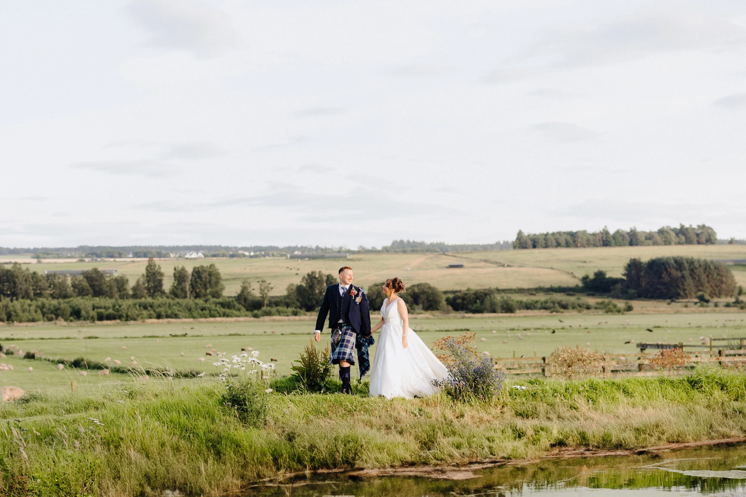 A couple in wedding attire walking together outdoors in a grassy field near a pond, with rolling hills and trees in the background.