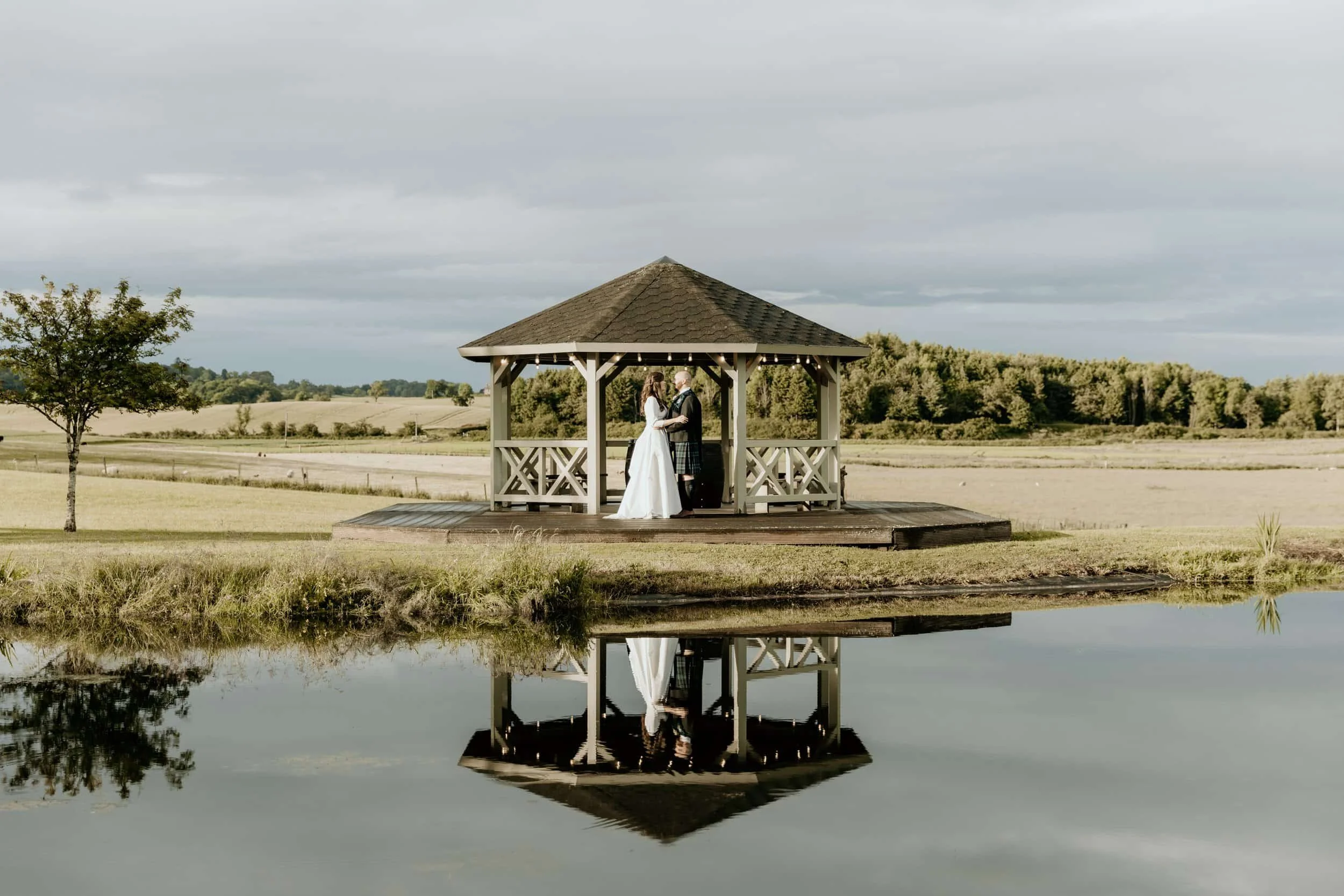 A couple in wedding attire standing under a gazebo by a pond, with their reflection visible in the water.
