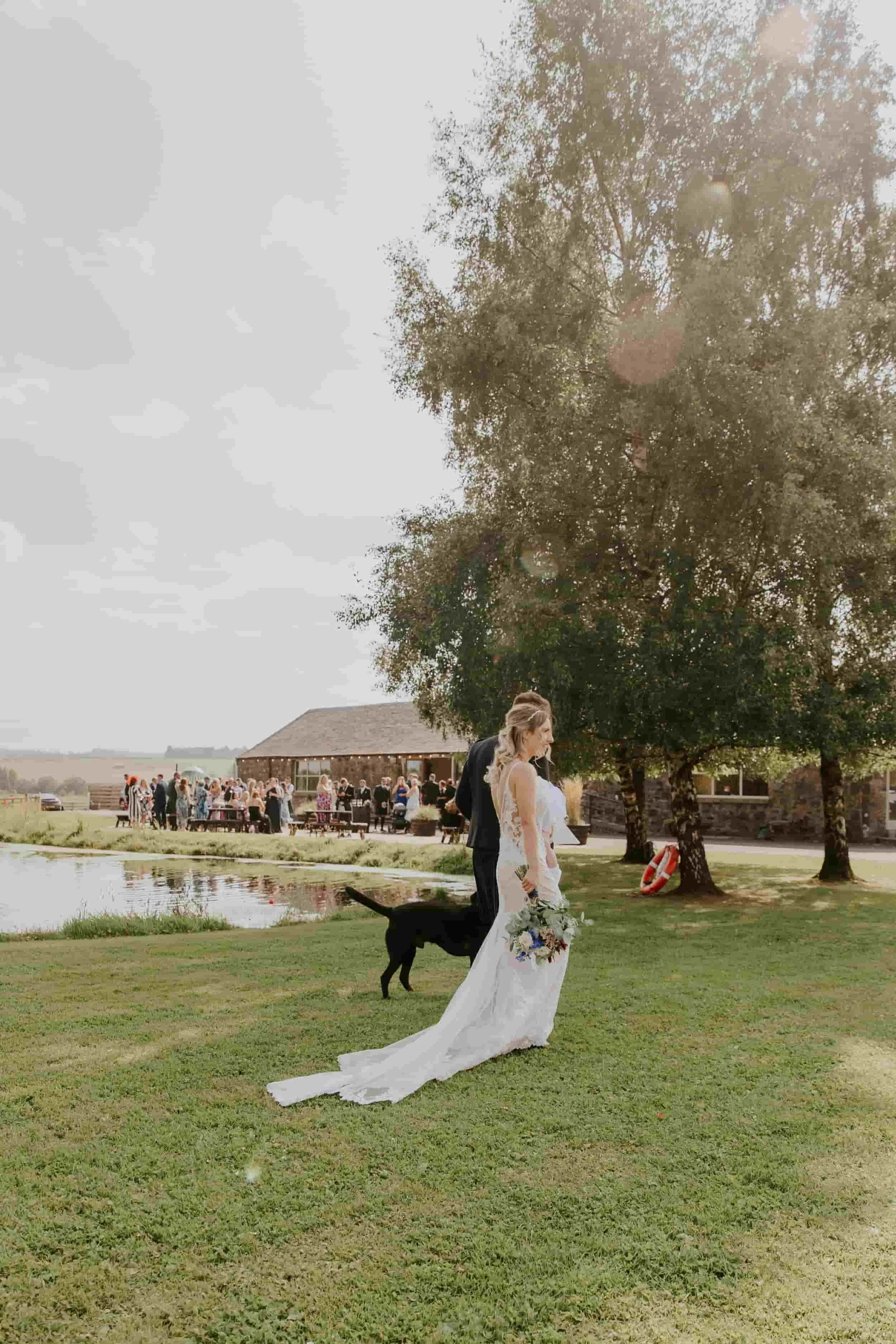 A bride and groom stand outdoors on a grassy area near a pond at a wedding reception. The bride is holding a bouquet and wearing a white wedding dress, while the groom is in a dark suit. A large black dog is standing nearby. In the background, guests