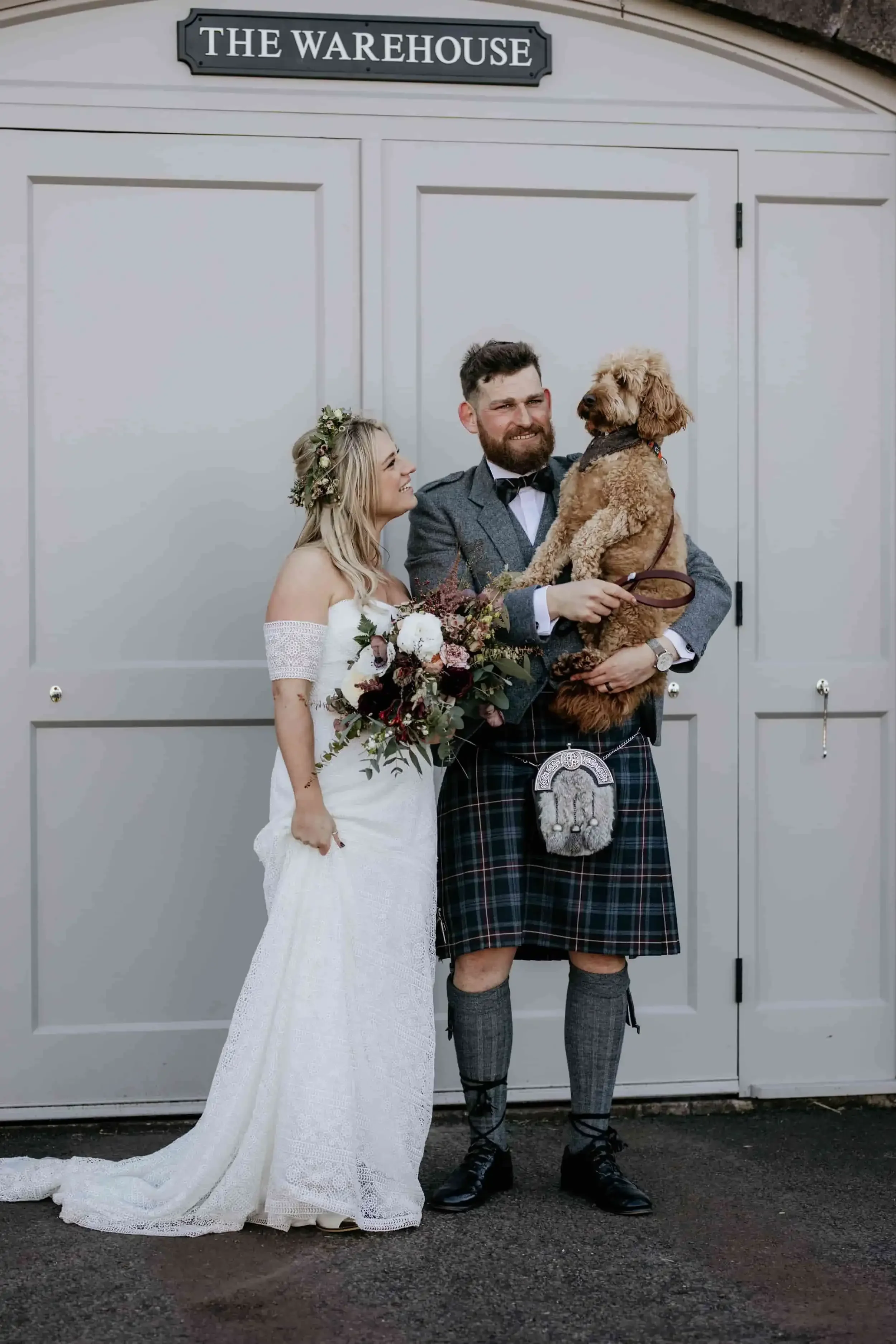 A bride and groom in wedding attire, with the groom holding a brown dog, standing in front of a gray door labeled 'The Warehouse'.