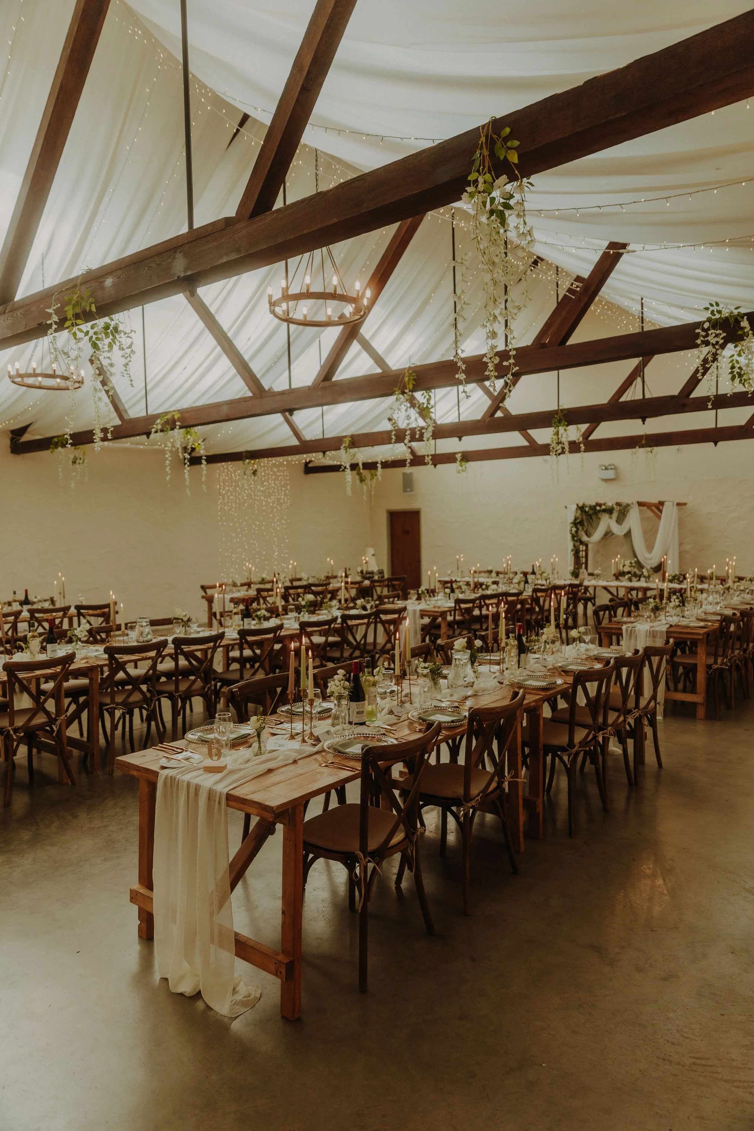 Elegant wedding reception setup with long wooden tables, candles, floral centerpieces, and string lights in a rustic hall with high ceiling and exposed wooden beams.