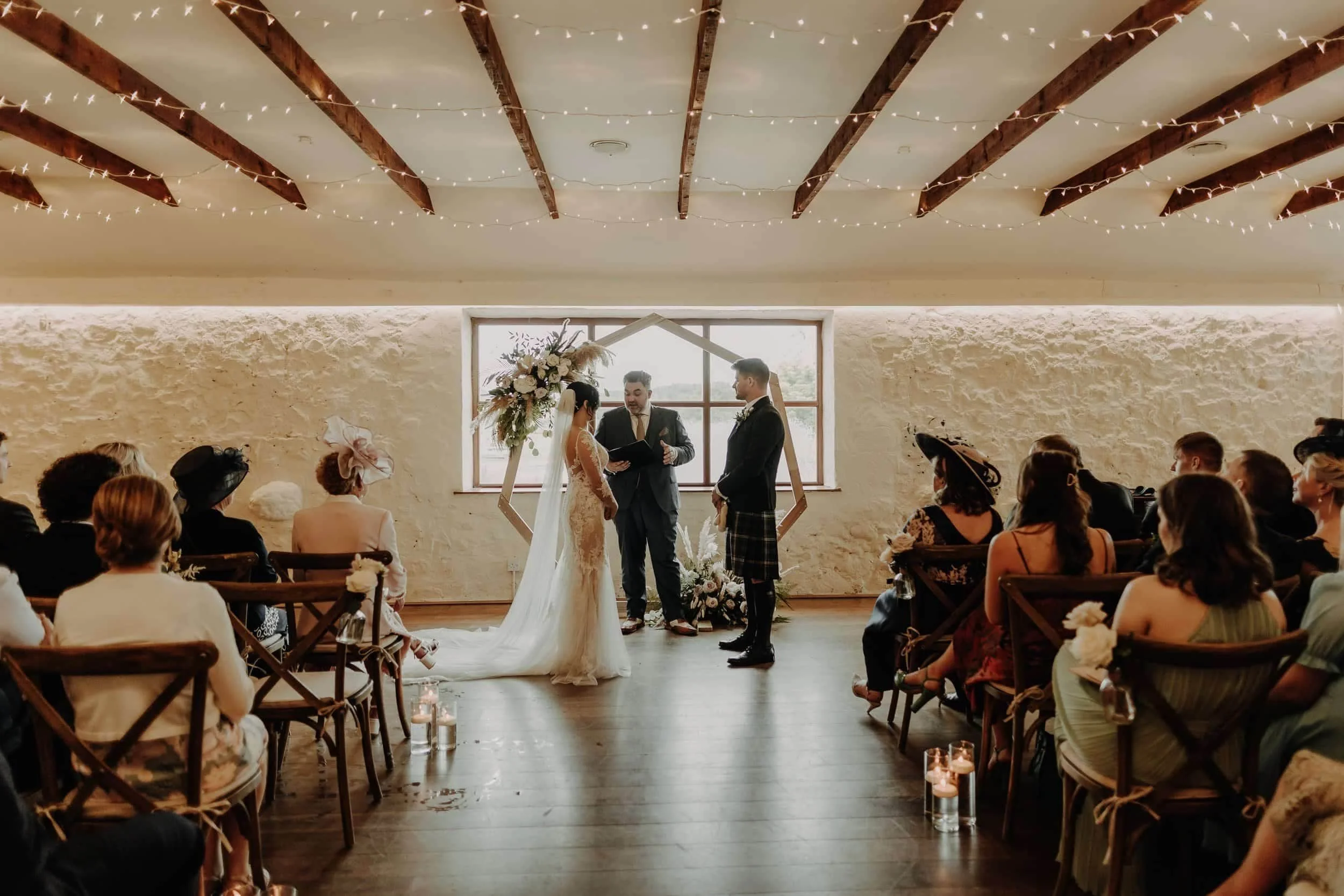 A wedding ceremony with a bride and groom standing before an officiant in a rustic indoor setting with exposed wooden beams, surrounded by seated guests in vintage and floral attire, during daytime with natural light from a large window.