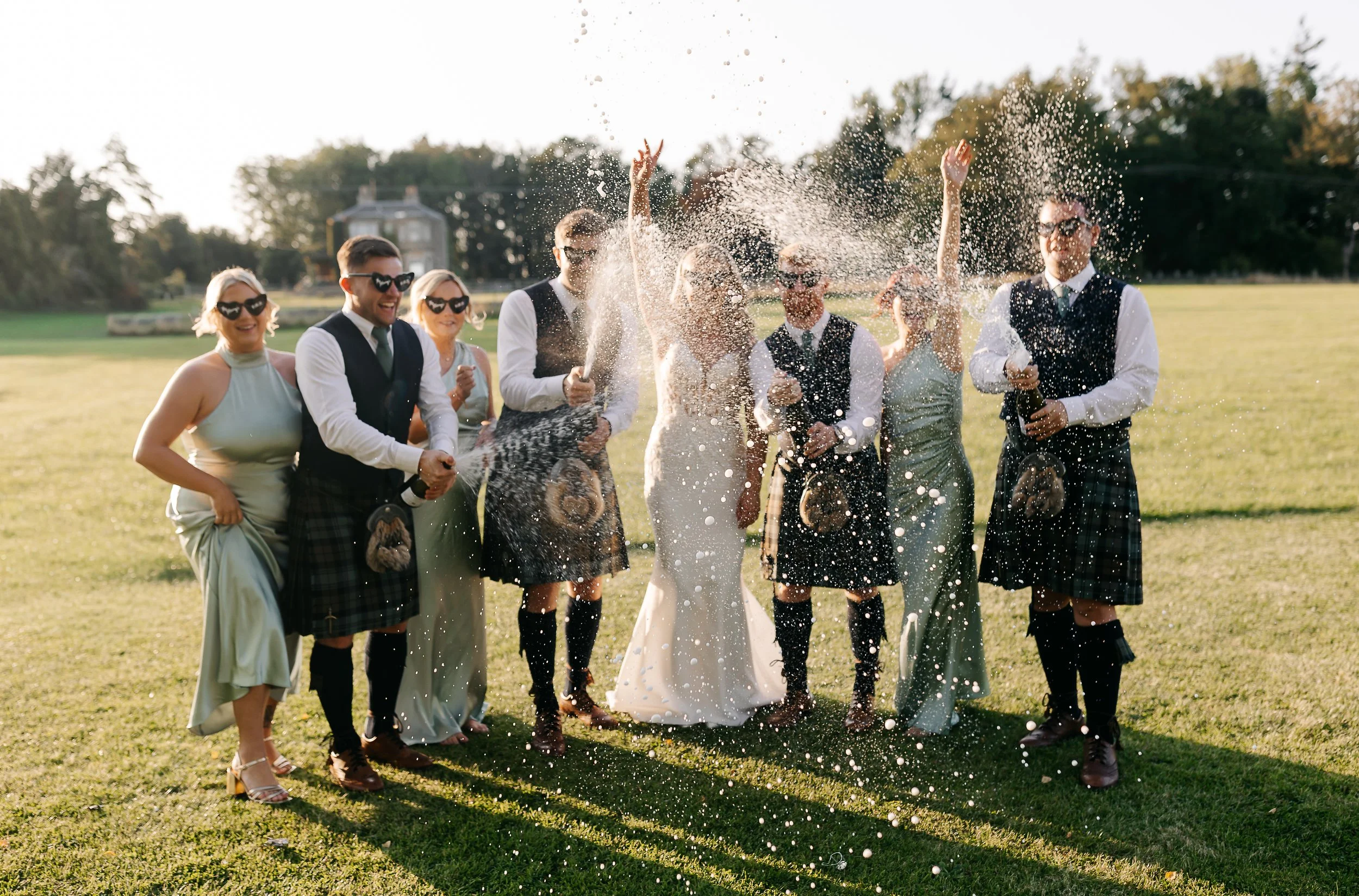 A group of people celebrating a wedding outdoors by spraying champagne, with the bride and groom in the center surrounded by friends in formal attire and sunglasses.
