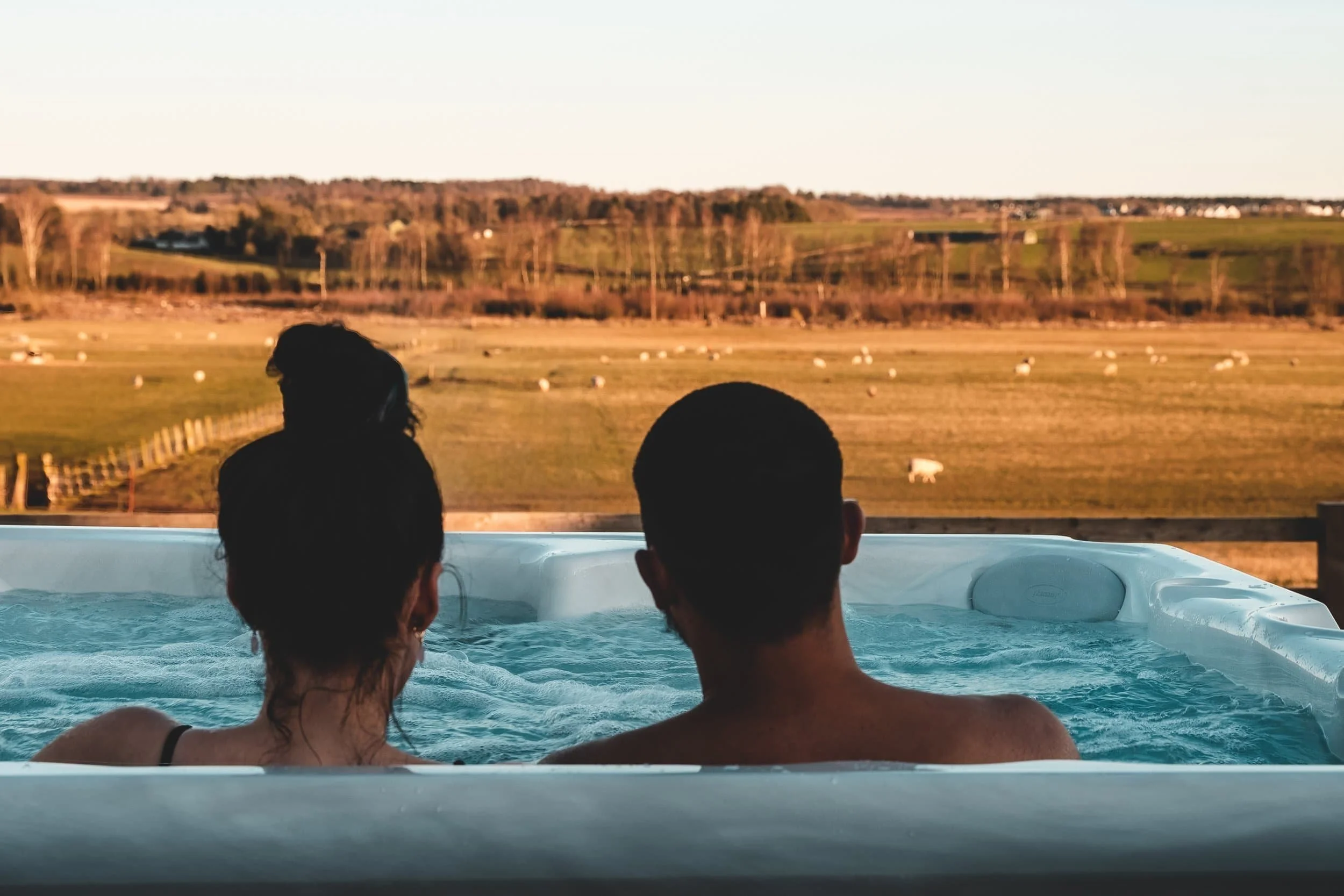 Two people sitting in a hot tub overlooking a countryside with green fields, sheep, trees, and distant houses at sunset.