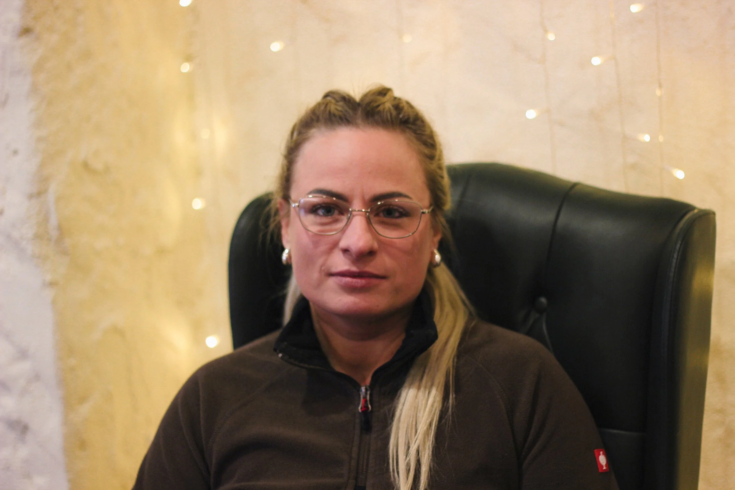 A woman with blonde hair, wearing glasses, earrings, and a dark zip-up jacket, sitting in a black leather chair with a golden and white background decorated with string lights.