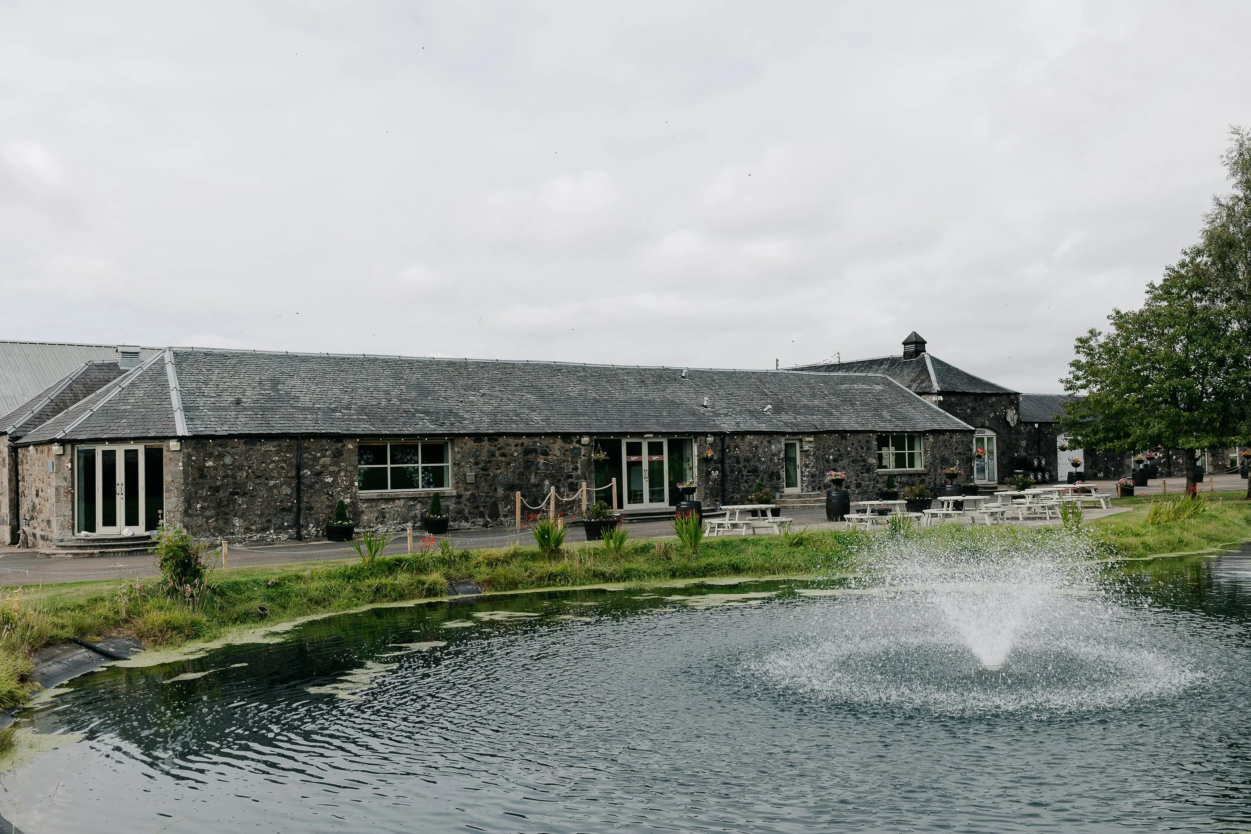 Stone building with outdoor seating near a pond with a fountain, trees, and cloudy sky.