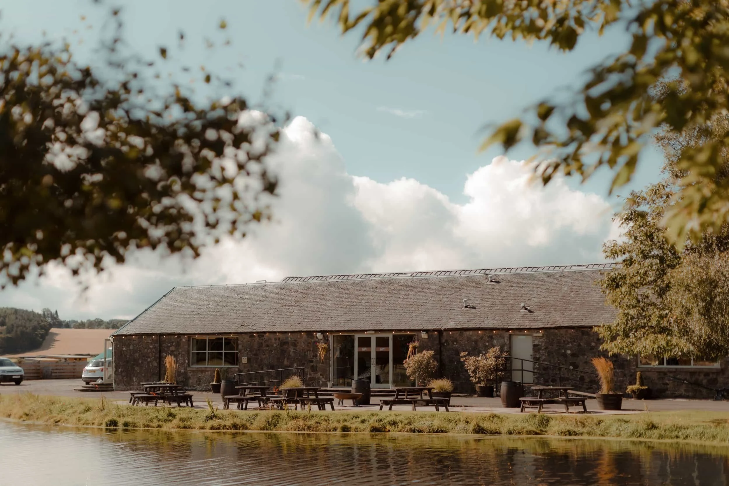 A rustic building with a sloped roof and large glass doors, surrounded by outdoor seating with potted plants, next to a body of water. Trees and a cloudy sky are in the background.