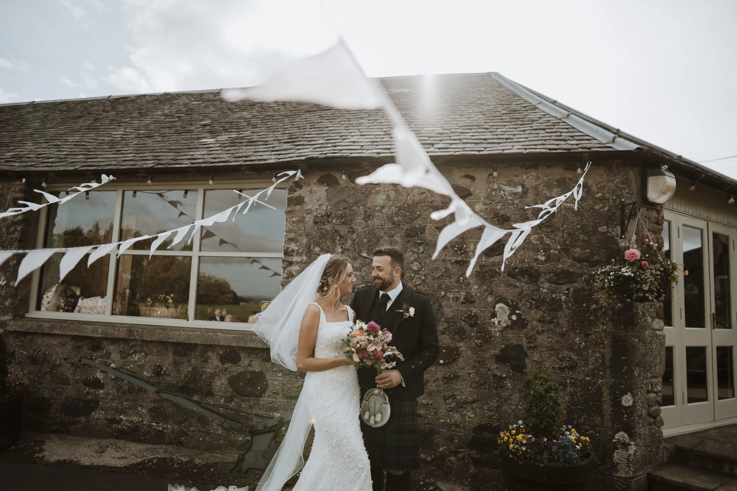 A bride and groom stand close together outdoors in front of a stone building decorated for a wedding. The bride wears a white wedding gown and veil, holding a bouquet of flowers, while the groom wears a black suit with a tartan kilt, holding a flower