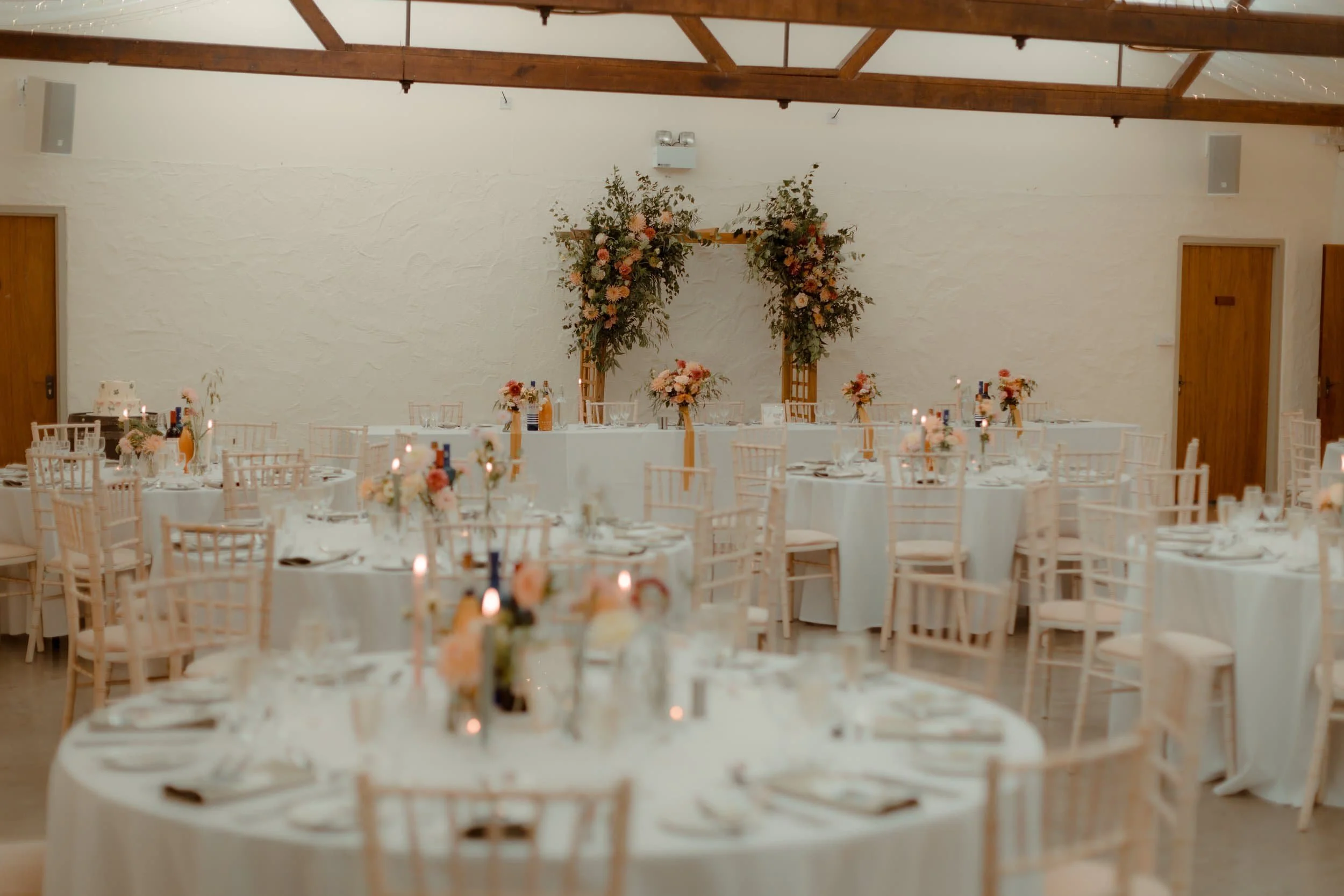 Wedding reception setup with round tables and white tablecloths, decorated with floral centerpieces and candles, and a floral arch backdrop.