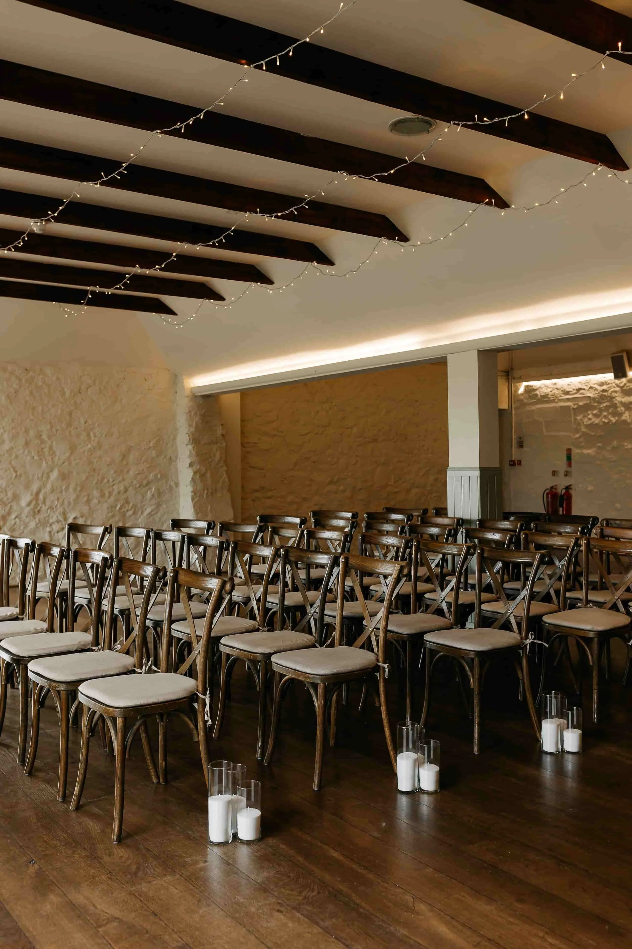 A decorated indoor event space with rows of wooden chairs and glass candle holders on the wooden floor. String lights are hanging from the ceiling, creating a warm ambiance.
