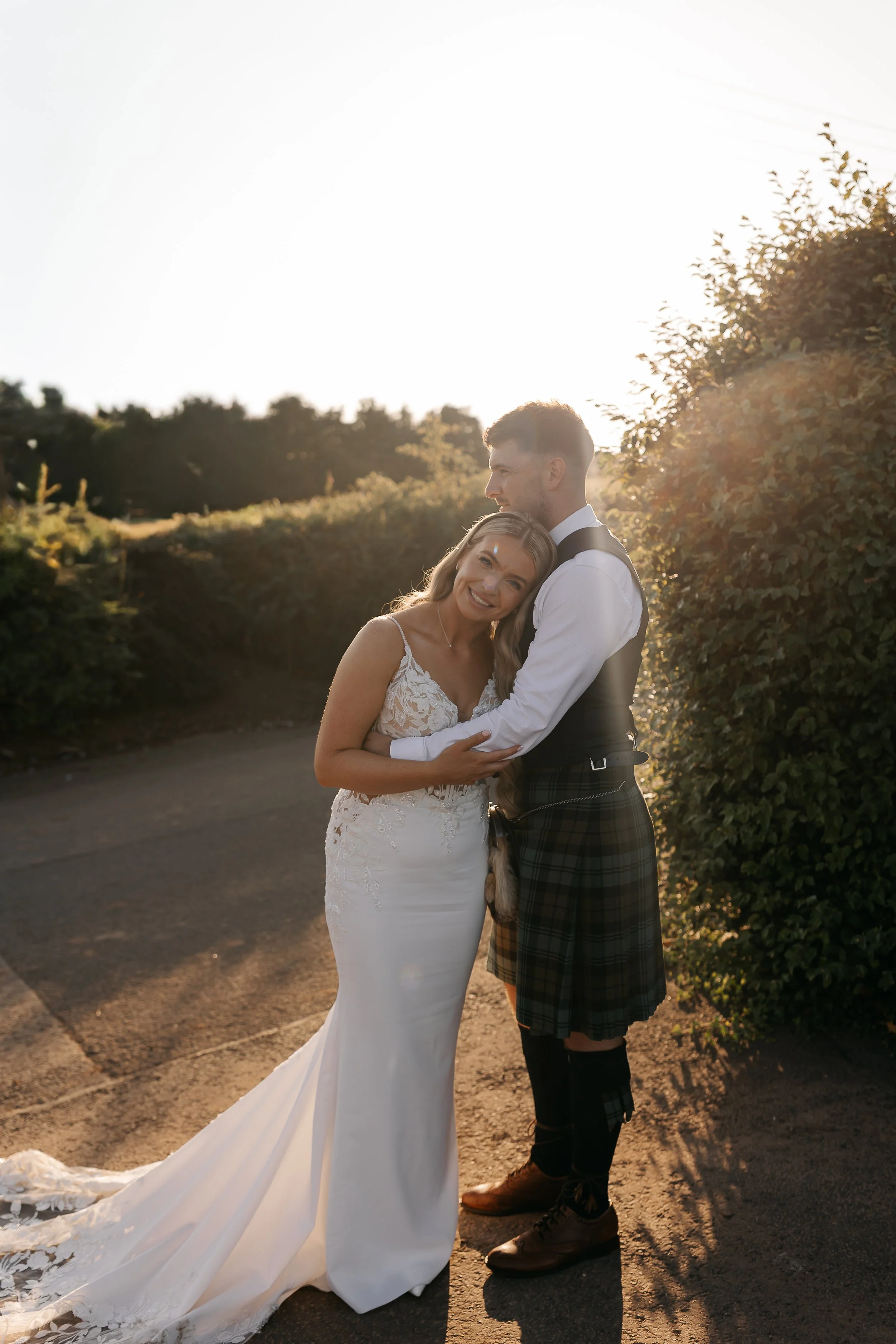 Bride and groom hugging outdoors at sunset, bride in white wedding dress with lace details, groom in formal attire with kilt, behind greenery and natural landscape.