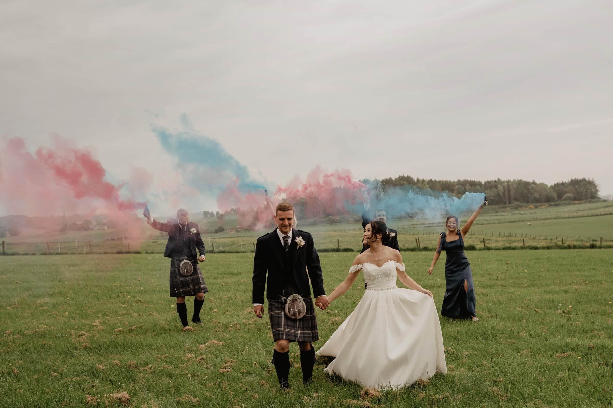 Bride and groom holding hands in a green field, with three friends behind them holding colorful smoke flares, during a wedding celebration.