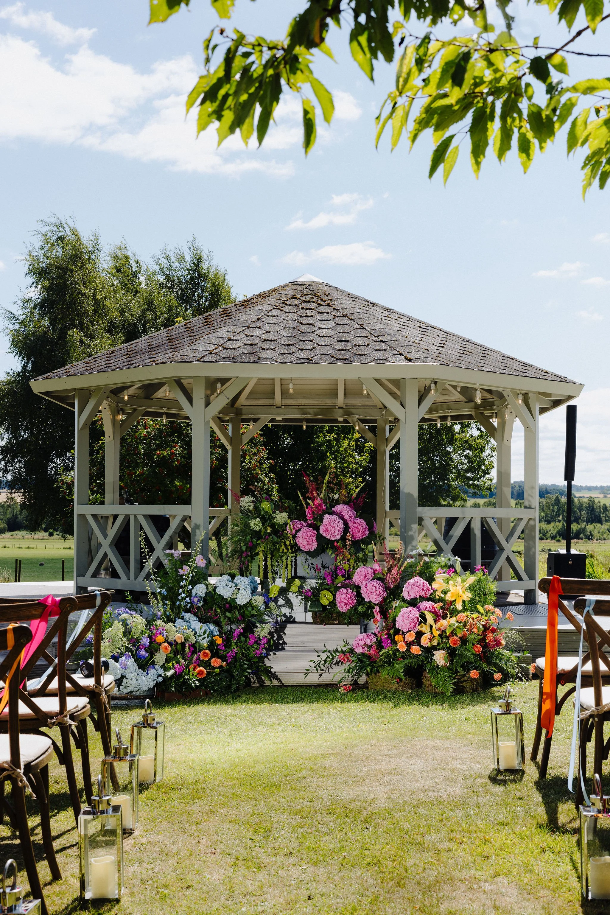 A decorated outdoor wedding gazebo with pink, white, and purple flowers, set up in a grassy field on a sunny day with scattered clouds.