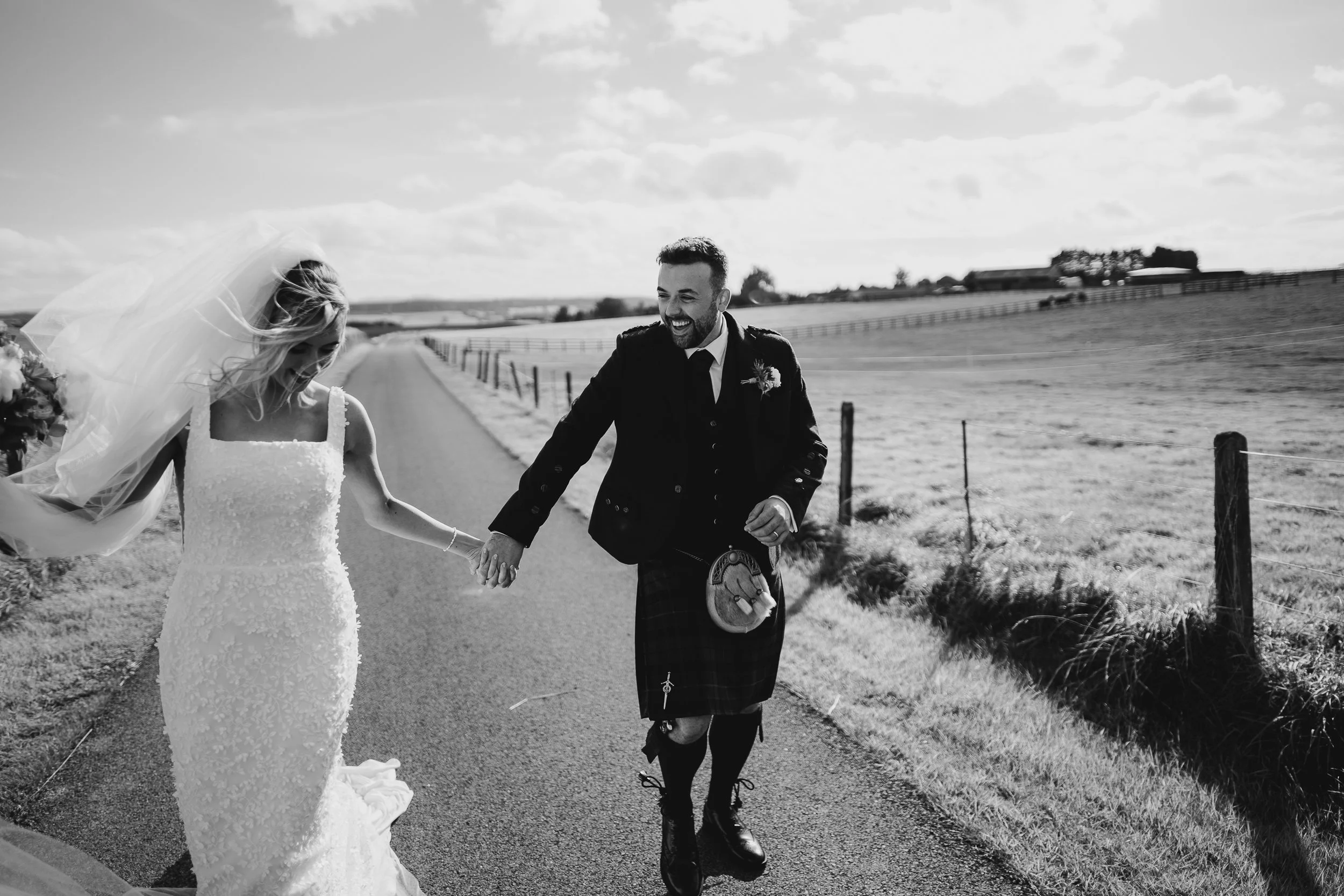 A bride and groom walking hand in hand along a country road, smiling and laughing outdoors on a sunny day.