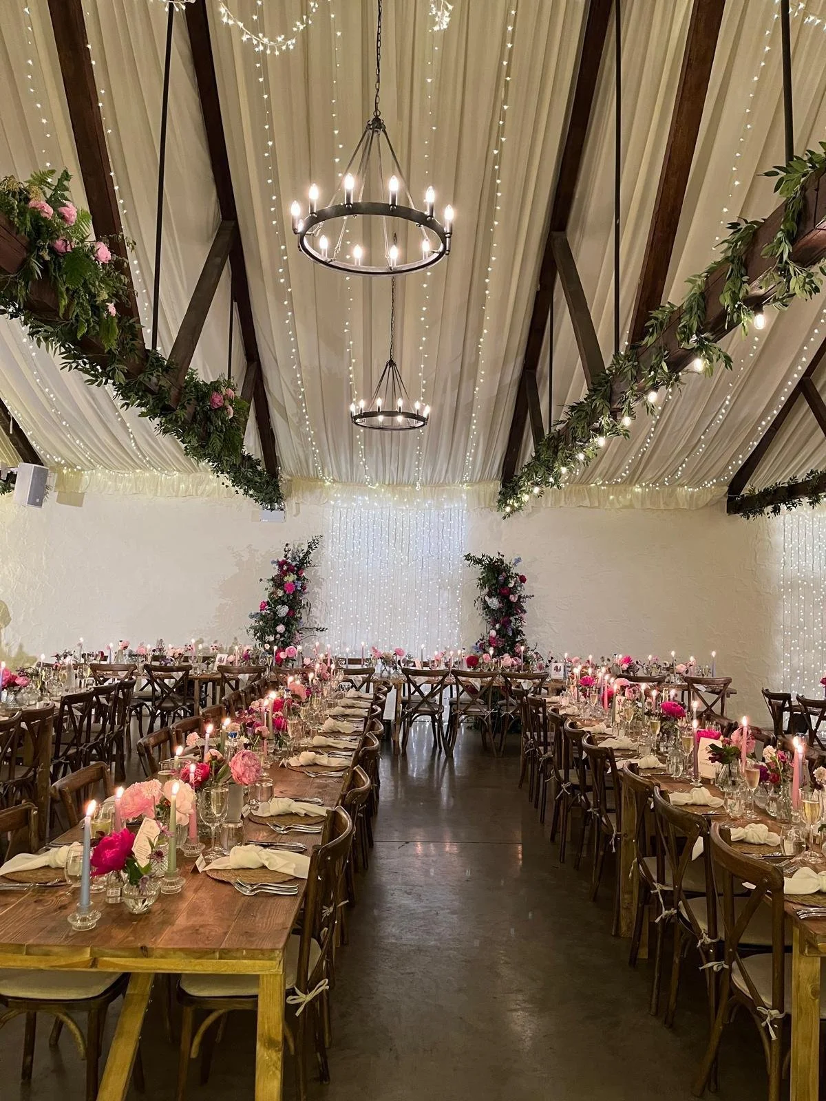Decorated banquet hall with long wooden tables set for a celebration, pink and white floral centerpieces, lit candles, draped white ceiling with string lights, and floral garlands hanging from walls.