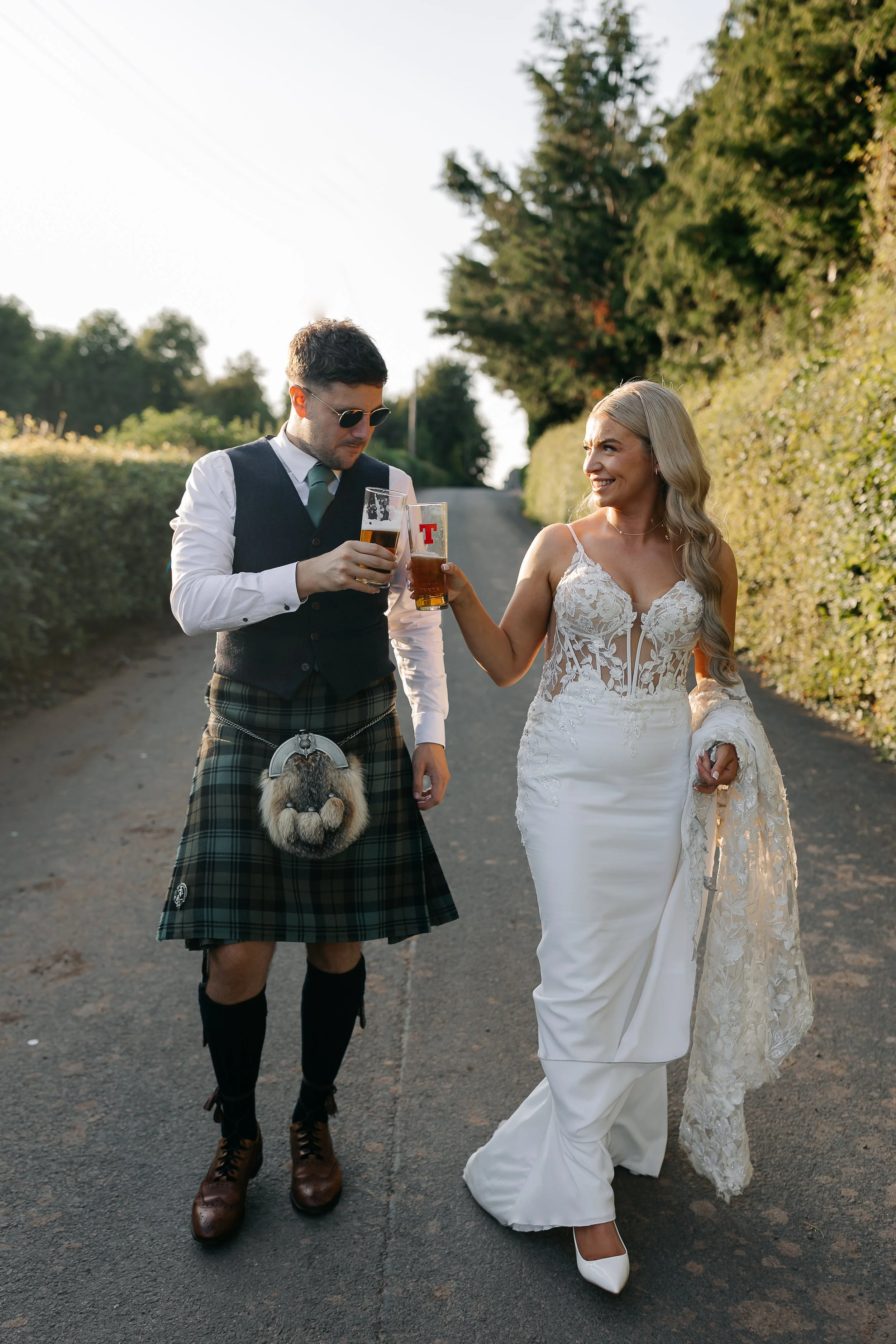 A couple dressed in wedding attire walking on a rural road, clinking glasses of beer, with the man wearing Scottish traditional clothing and the woman in a white wedding gown.