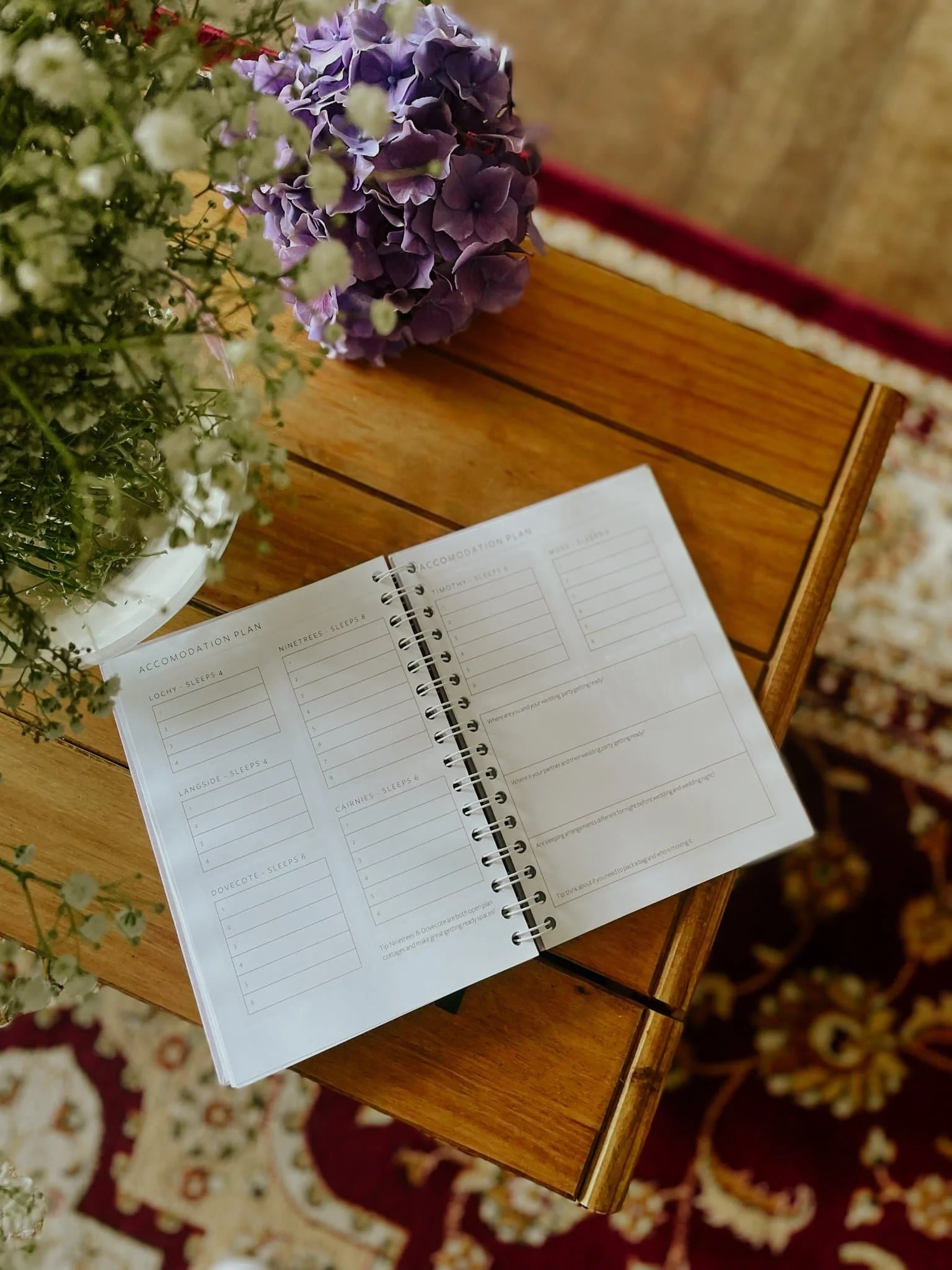 Open spiral-bound planner or journal on a wooden table with sections for accommodation plans, surrounded by purple hydrangeas and white baby's breath flowers, with a patterned carpet underneath.