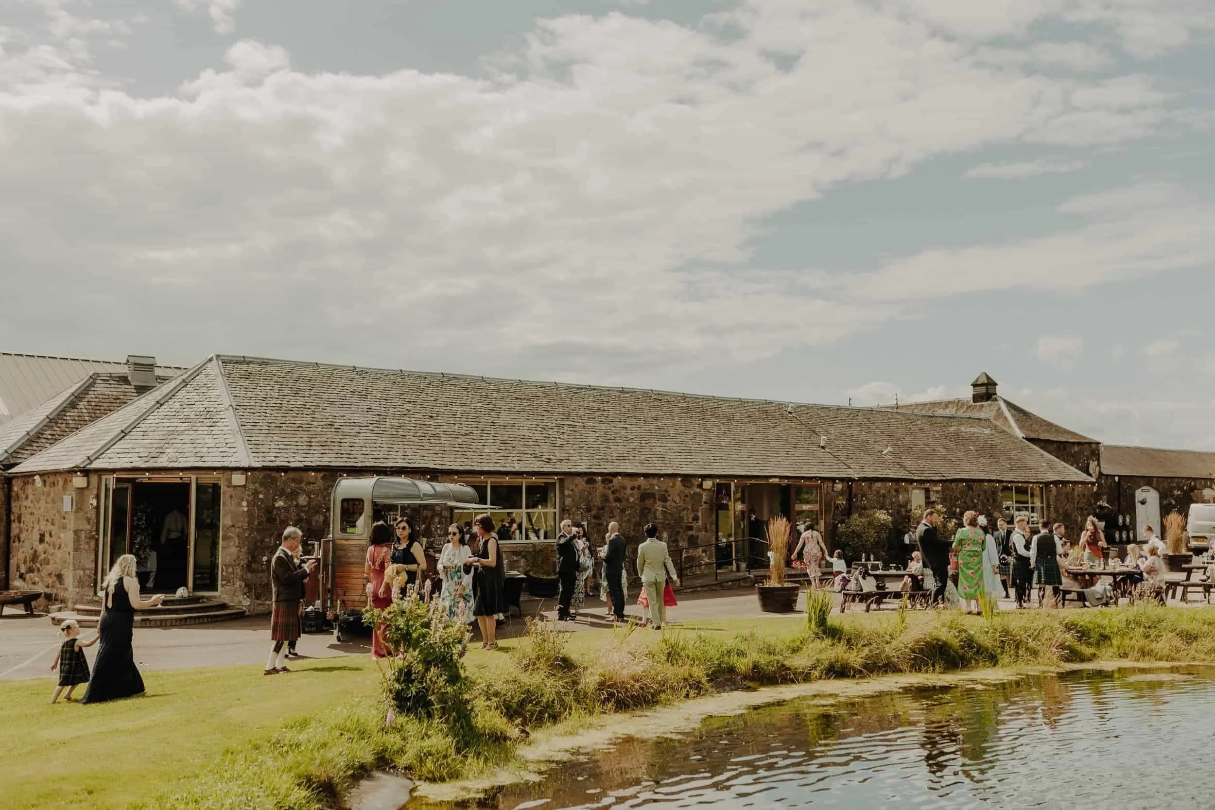 People gather outside a rustic stone building near a pond under a partly cloudy sky, dressed for a social or festive event.