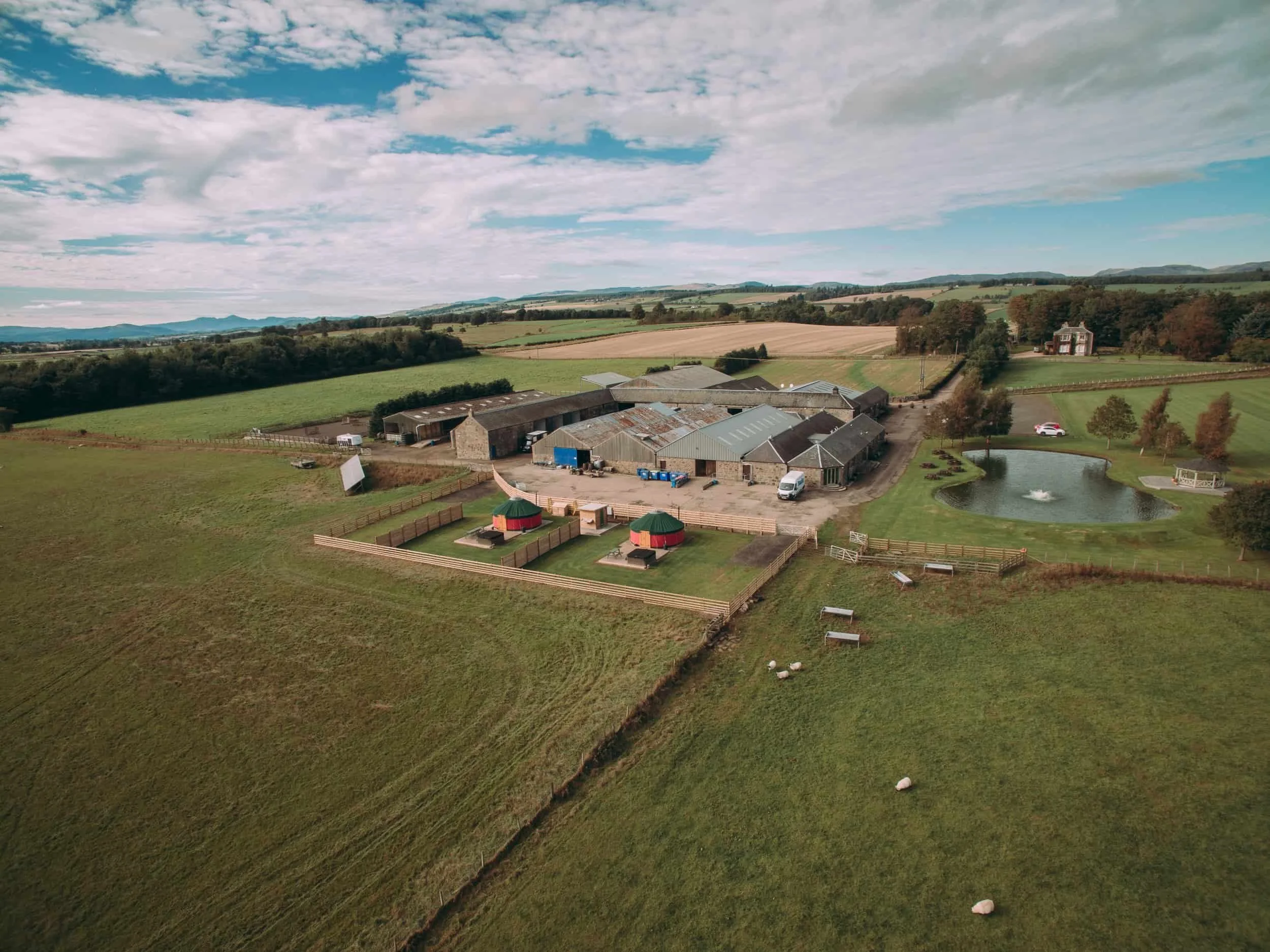 Aerial view of a farm with barns, green fields, a small pond, and scattered sheep under a partly cloudy sky.