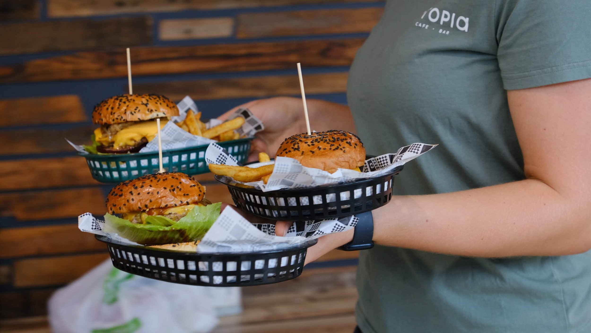 Person holding three baskets of cheeseburgers and fries in a restaurant with wooden wall decor.