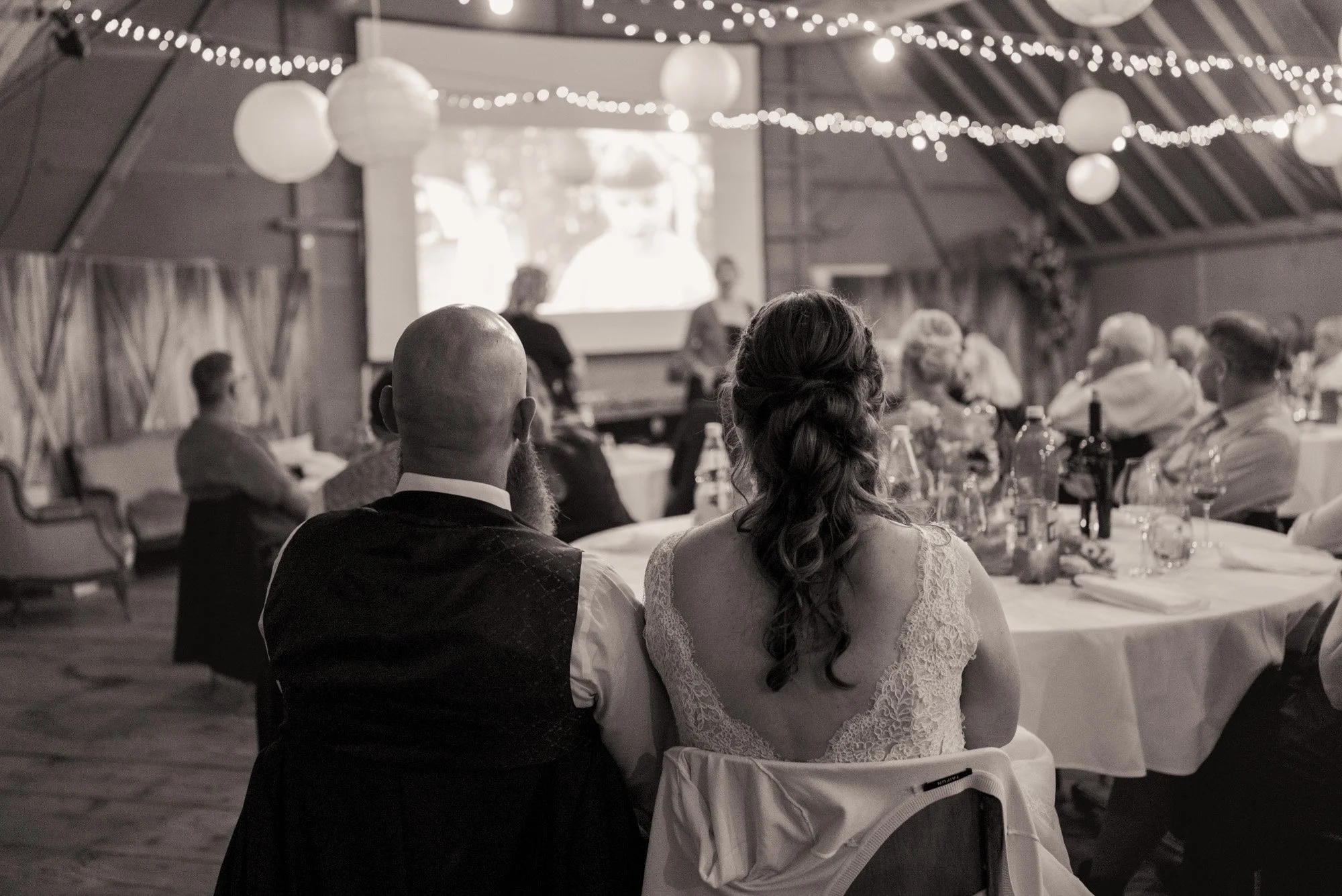 Paare sitzen bei einer Hochzeit im Innenraum eines Festsaals, schauen auf eine Leinwand mit einer Videoaufnahme, umgeben von Dekoration und Lichterketten.