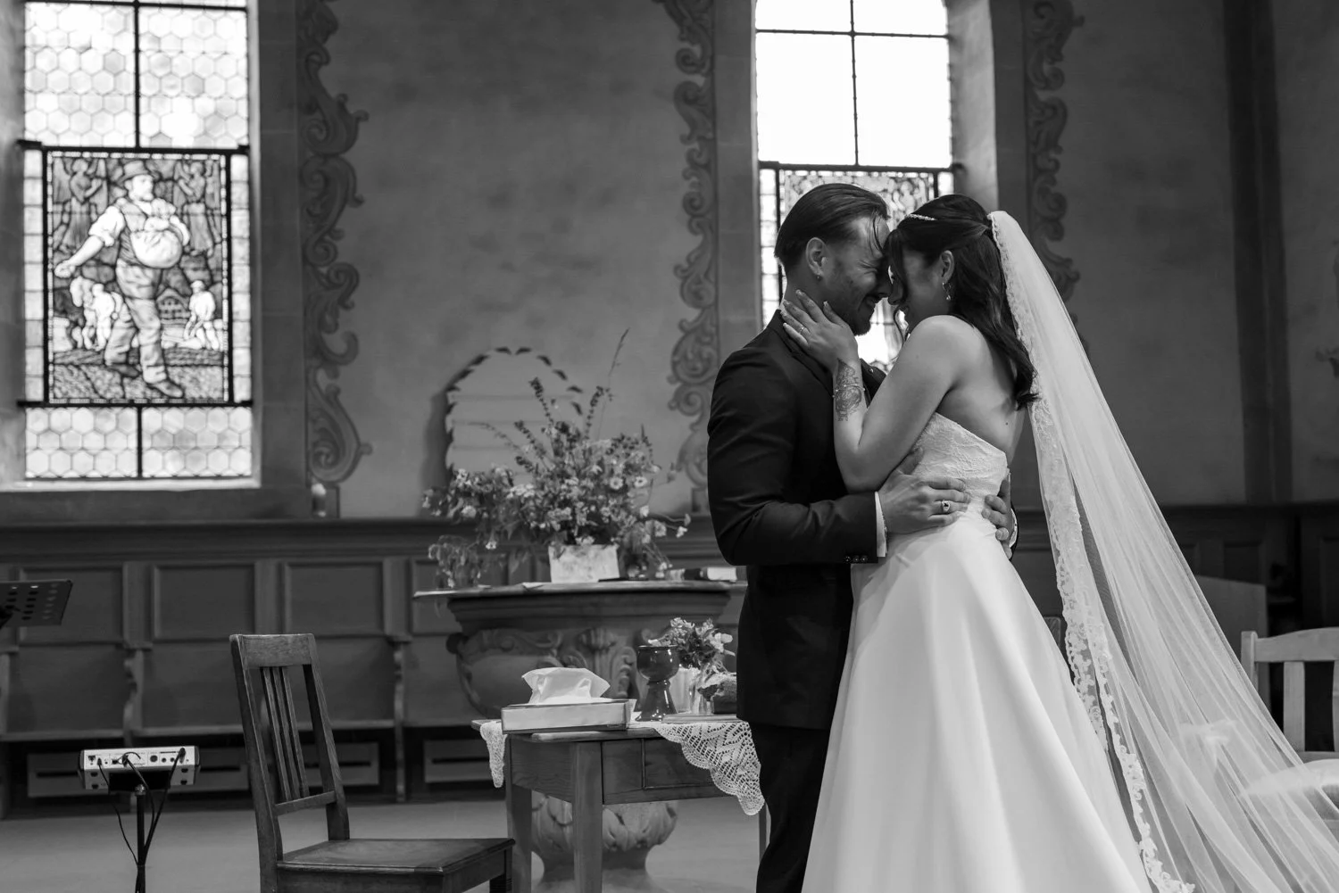 Ein schwarz-weiß Foto eines Brautpaares bei ihrer Trauung in einer Kirche, sie stehen sich nah, berühren ihre Stirnen, die Frau trägt ein langes Brautkleid mit Schleier, der Mann einen schwarzen Anzug.