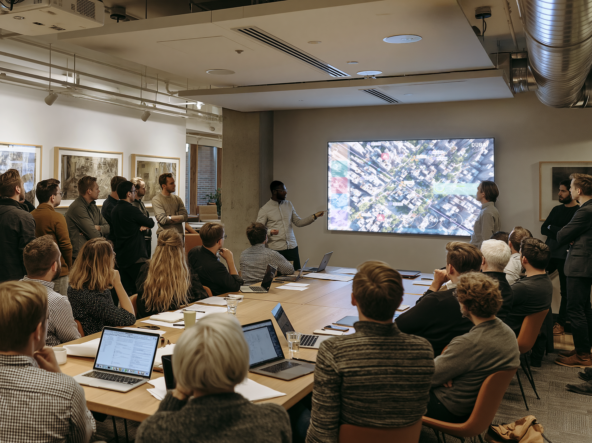 A group of people in a conference room attending a presentation. A man in a white shirt is speaking while pointing at a large screen displaying a digital twin with data overlays. Others are seated around a long table, some using laptops.