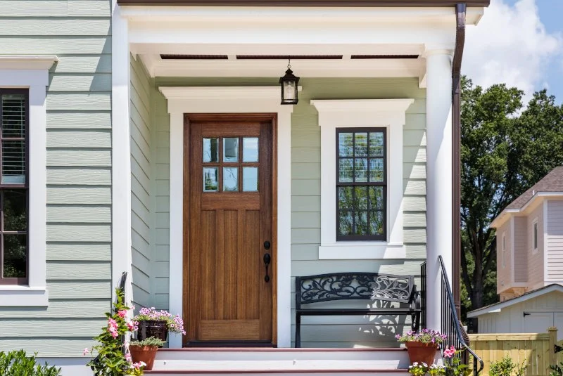 Front porch with wooden door, green siding, black bench, potted flowers, and overhead lantern.