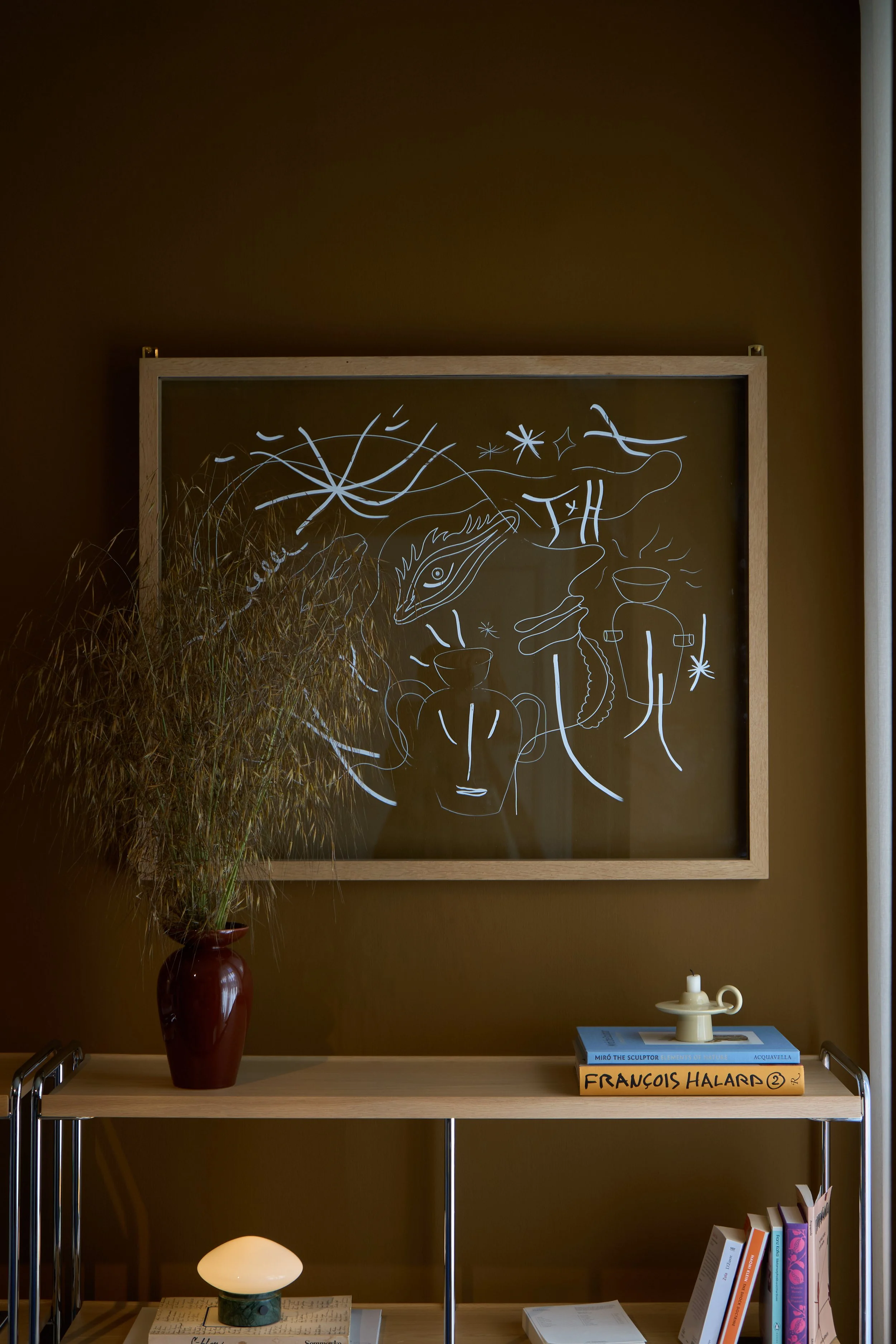 Interior photography captured inside the &Tradition Townhouse Apartment in Copenhagen. The frame documents a moody, tobacco-brown gallery wall featuring a modern chrome console shelf styled with an eclectic mix of abstract graphic art, design books, 