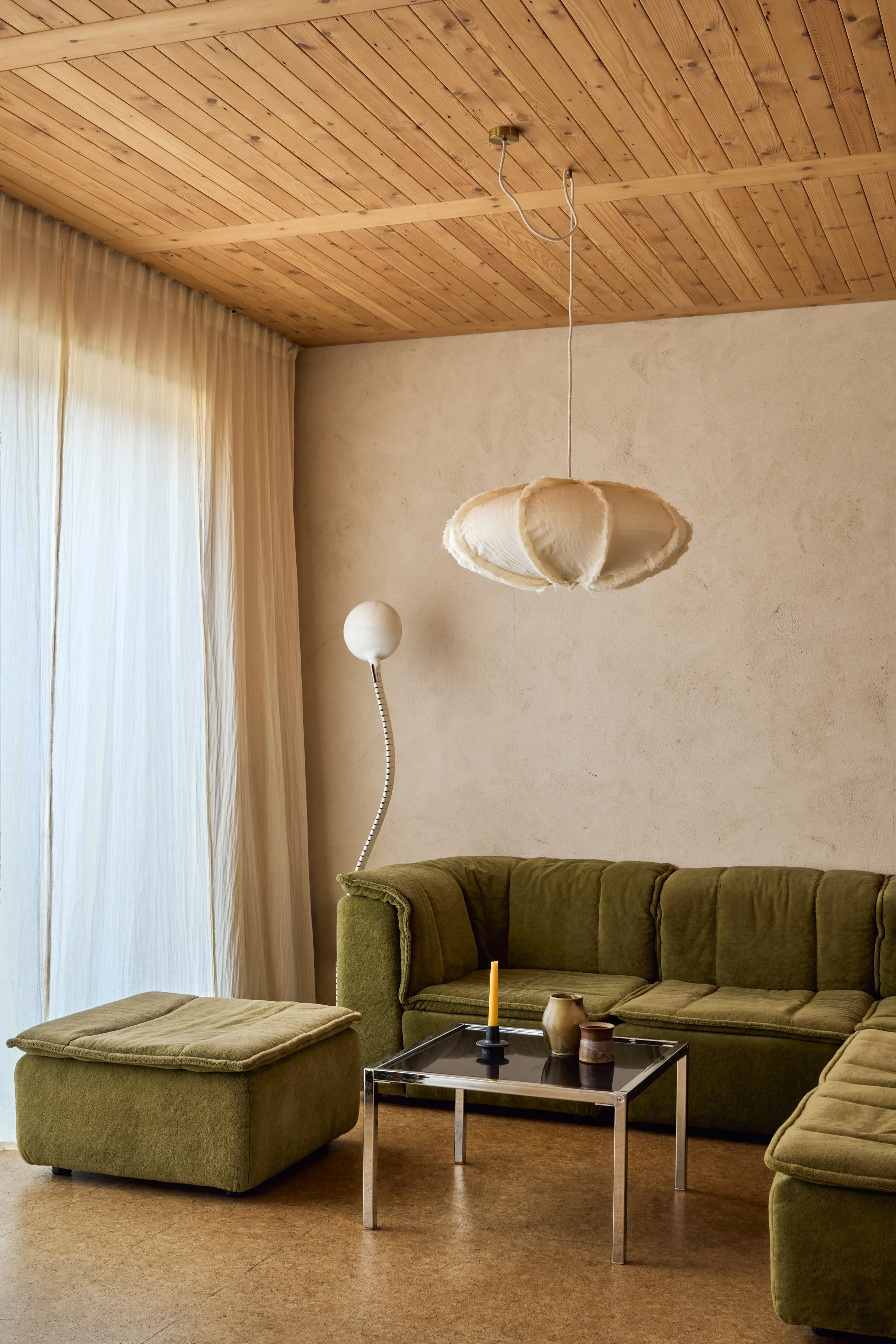 Warm minimalist living room in the award-winning Nina's House retrofit featuring an olive green modular velvet sofa, a slatted timber ceiling, and a large linen pendant light. 