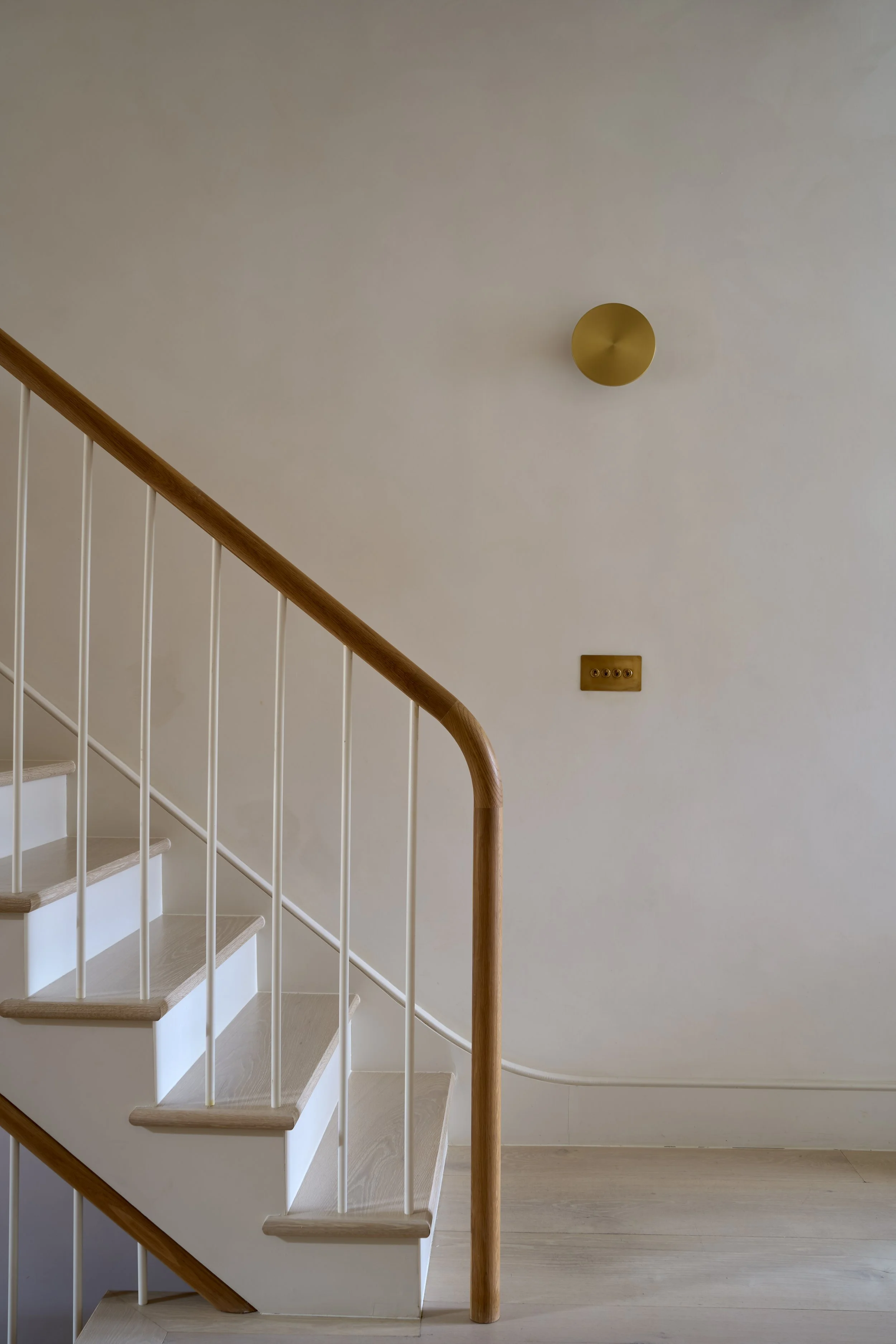 Architectural photograph of a minimalist modern staircase featuring light oak wood treads, a curved oak handrail, slim white balusters, and a brass wall sconce light fixture against a plaster wall.