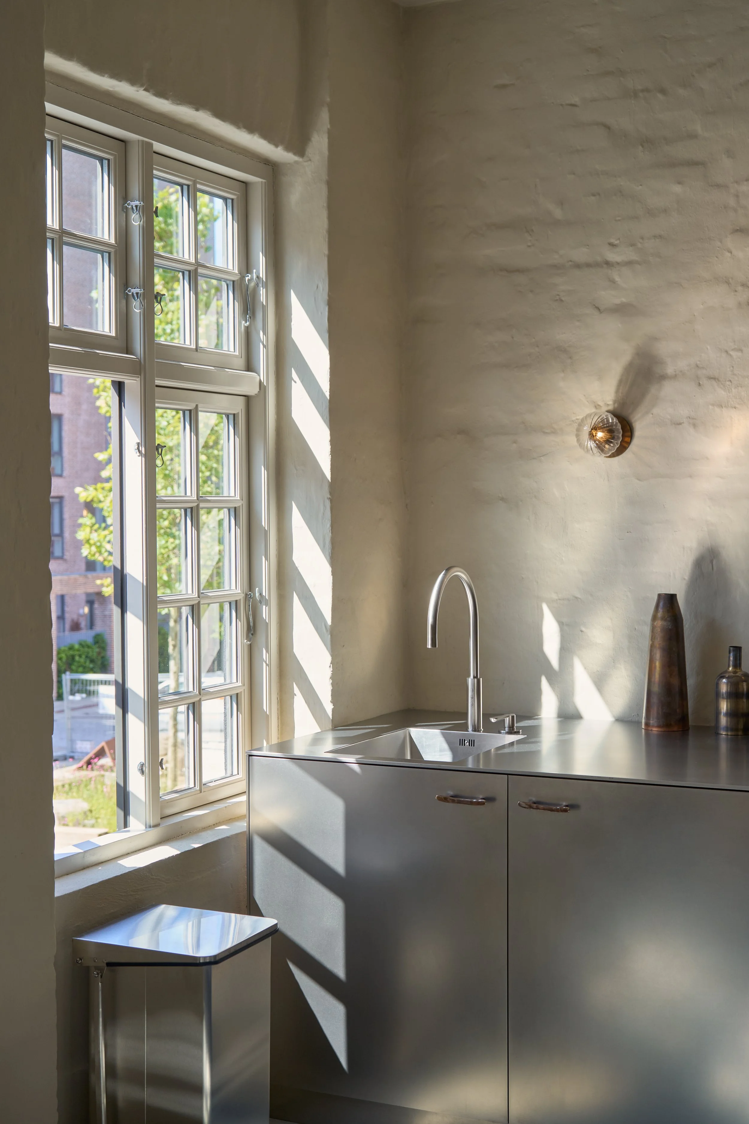 Architectural photograph of a minimalist kitchen area at the St. Leo residence in Copenhagen, featuring sleek stainless steel cabinets and countertop under dramatic window sunlight streaming across a textured plaster wall.