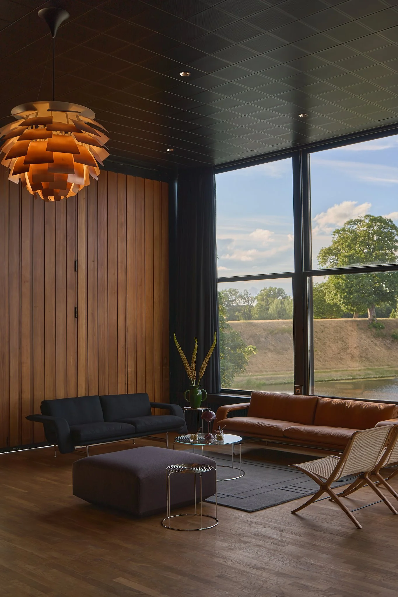 Moody interior photography of a modernist living space featuring an iconic Louis Poulsen Artichoke pendant light and leather sofas by &Tradition. Shot with natural available light to highlight the honest materiality of timber walls, clean structural 