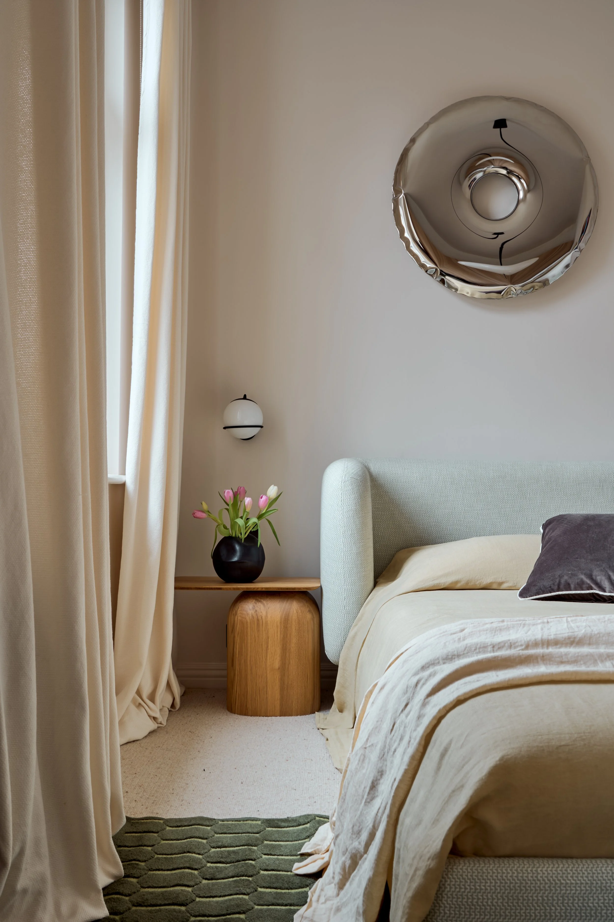 Editorial bedroom interior photograph showcasing warm minimalism and tactile materiality. The composition captures a modern mint-green upholstered bed with natural linen bedding, a sculptural oak bedside table styled with fresh tulips, and a reflecti