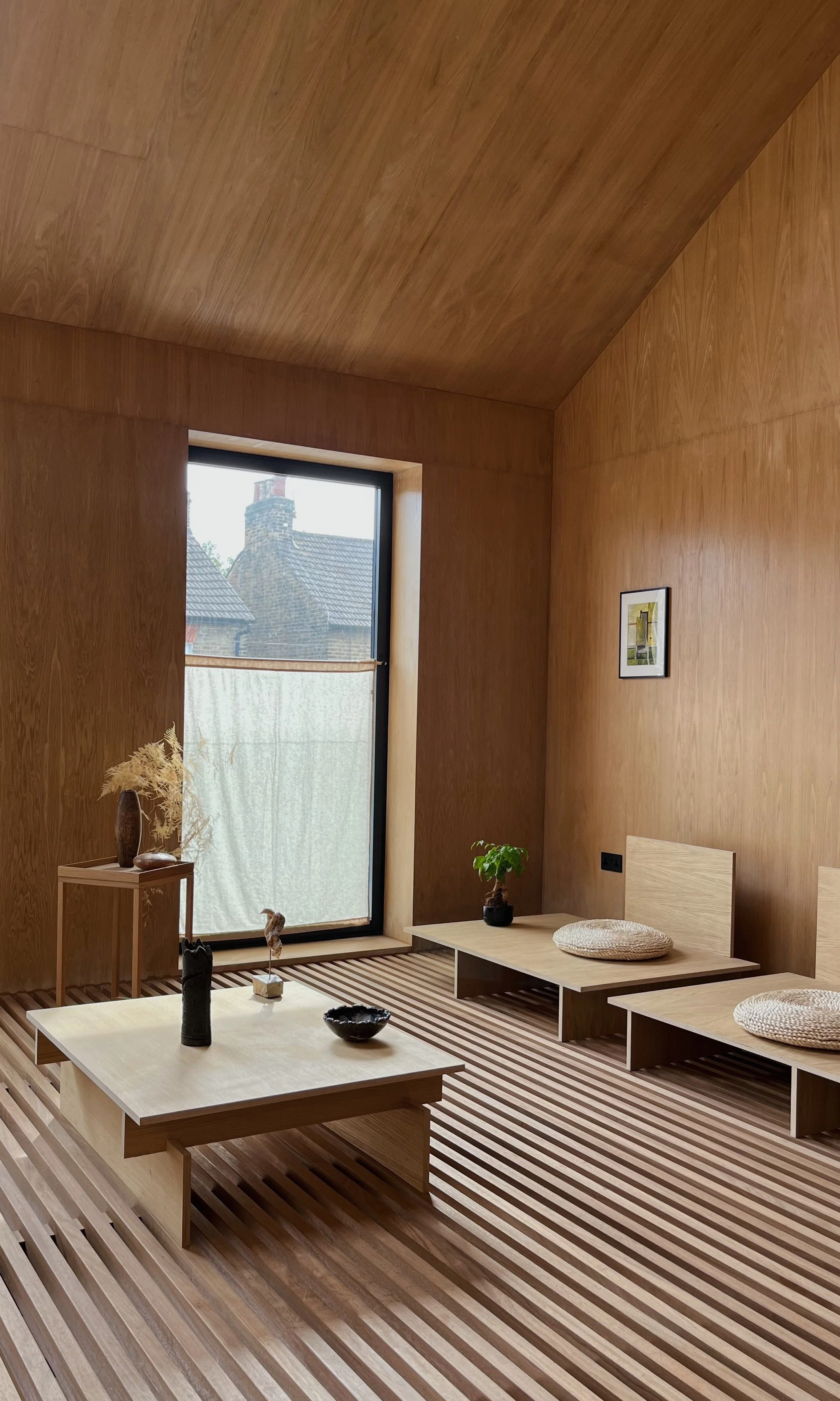 Minimalist living room inside Cooper House featuring uniform natural wood paneling, a distinctive slatted iroko wooden slatted floor, a low profile coffee table with ceramic decor, and a tall vertical window framing a London neighborhood view.