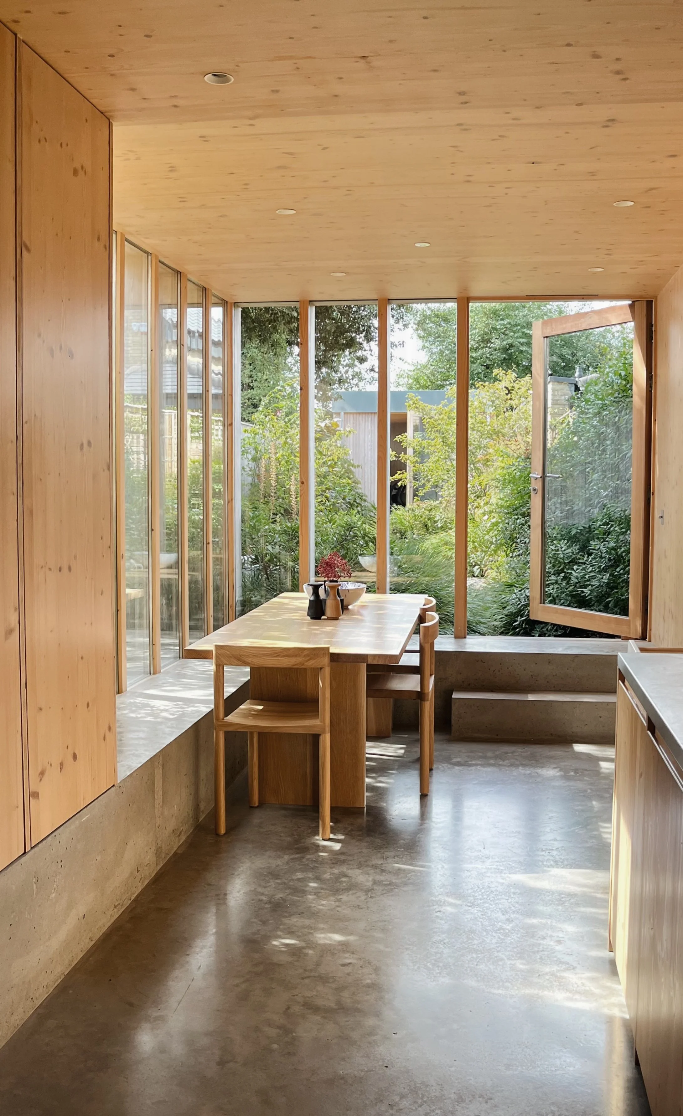Modern residential kitchen and dining extension with a seamless indoor-outdoor flow, highlighting cross-laminated timber (CLT) paneling, built-in concrete seating bench, minimalist oak chairs, and a pivoted timber glass door opening to a lush garden.