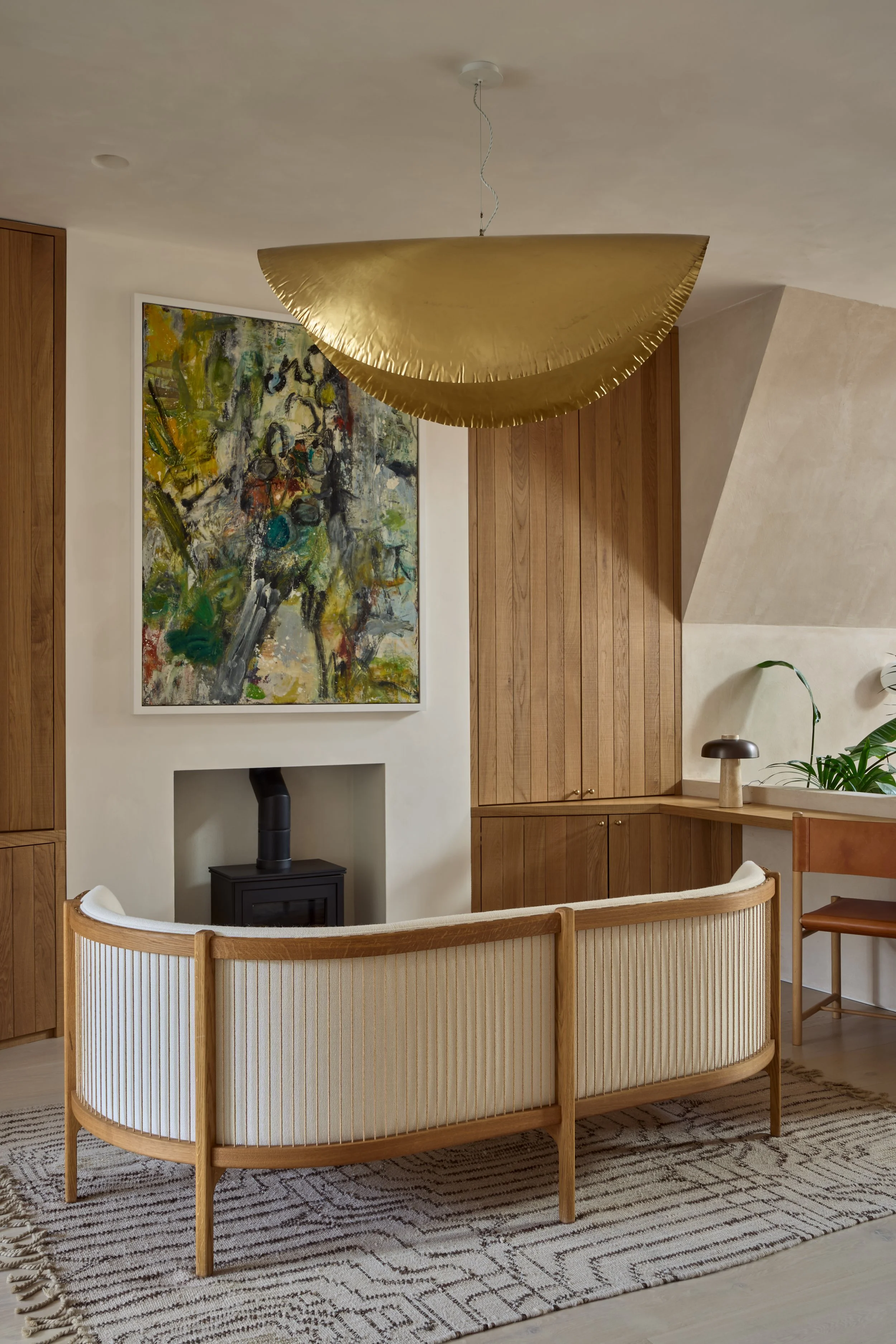 Professional interior photograph of a modern living room featuring a curved Carl Hansen & Søn Sideways Sofa, a large abstract painting, a black wood-burning stove, and a statement gold pendant light fixture.