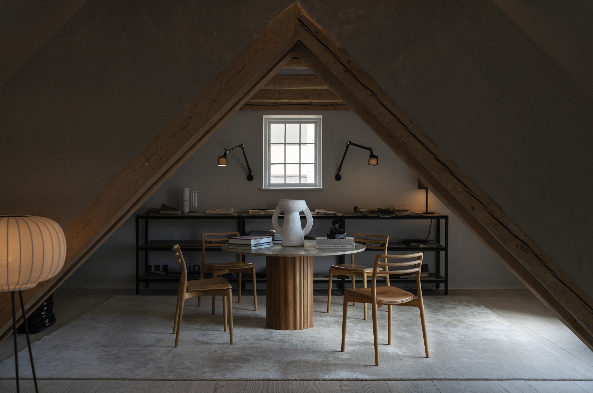 Minimalist attic workspace in the Vipp Loft with exposed wooden A-frame beams, featuring a round fluted wood table, light oak Vipp Cabin Chairs, and a large white Serax decorative vase. 